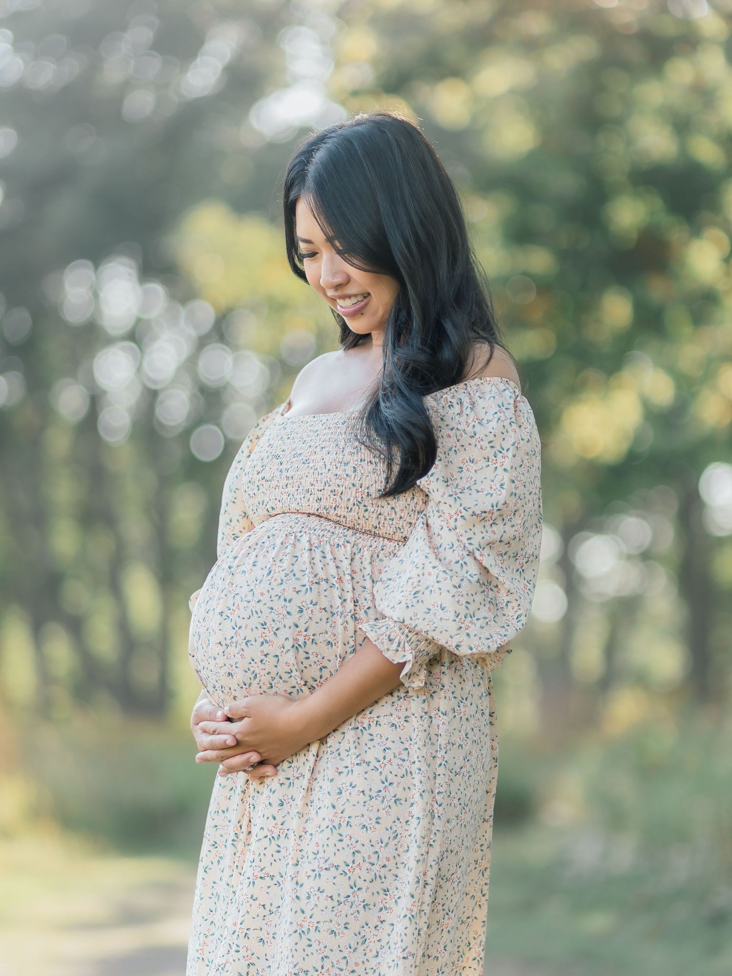 Expectant mother in a floral dress stands outdoors in a dreamy sunlit forest setting during a maternity photoshoot.