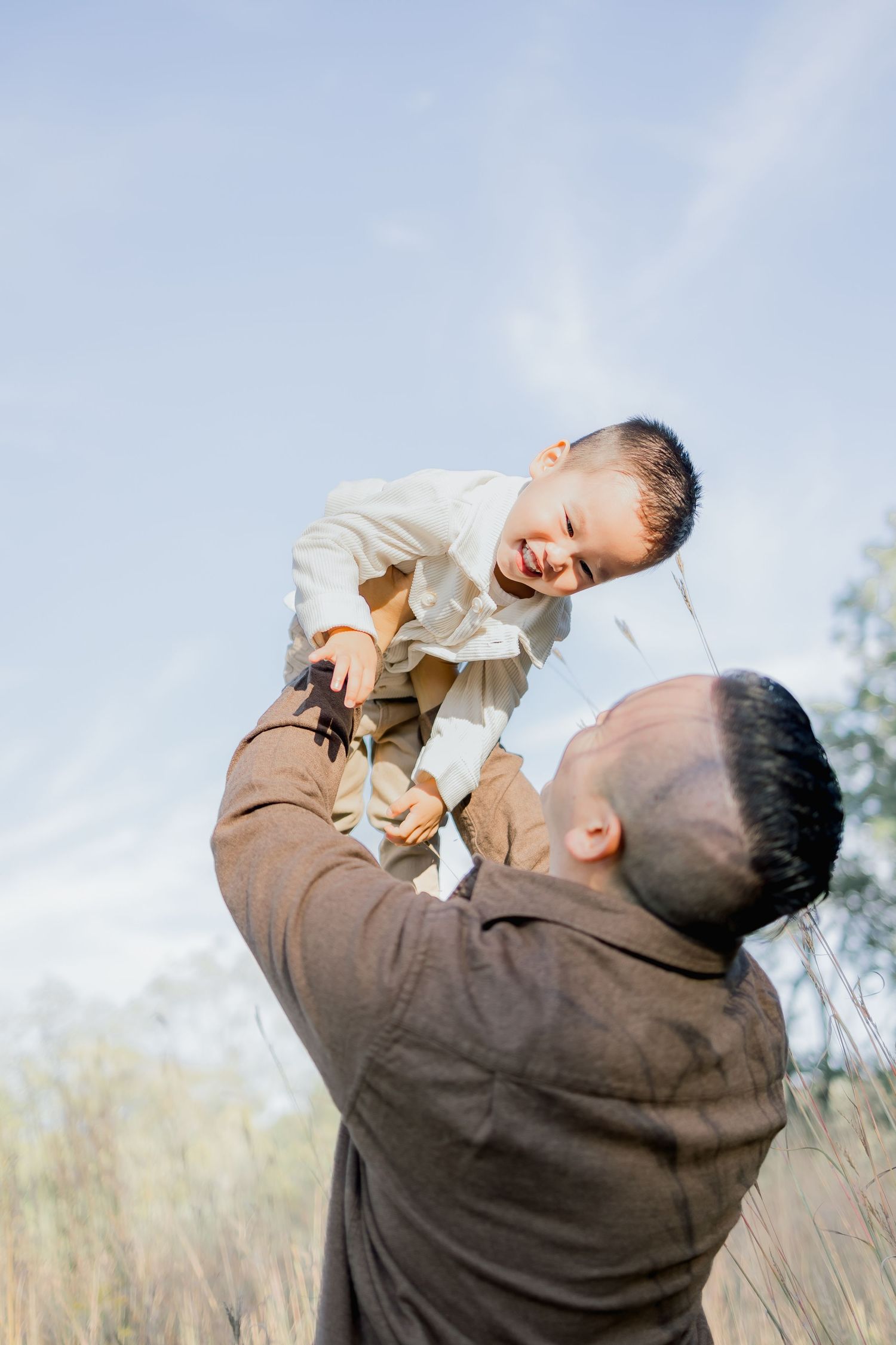 A parent wearing a brown sweater lifts and plays with a small child against a bright outdoor background.