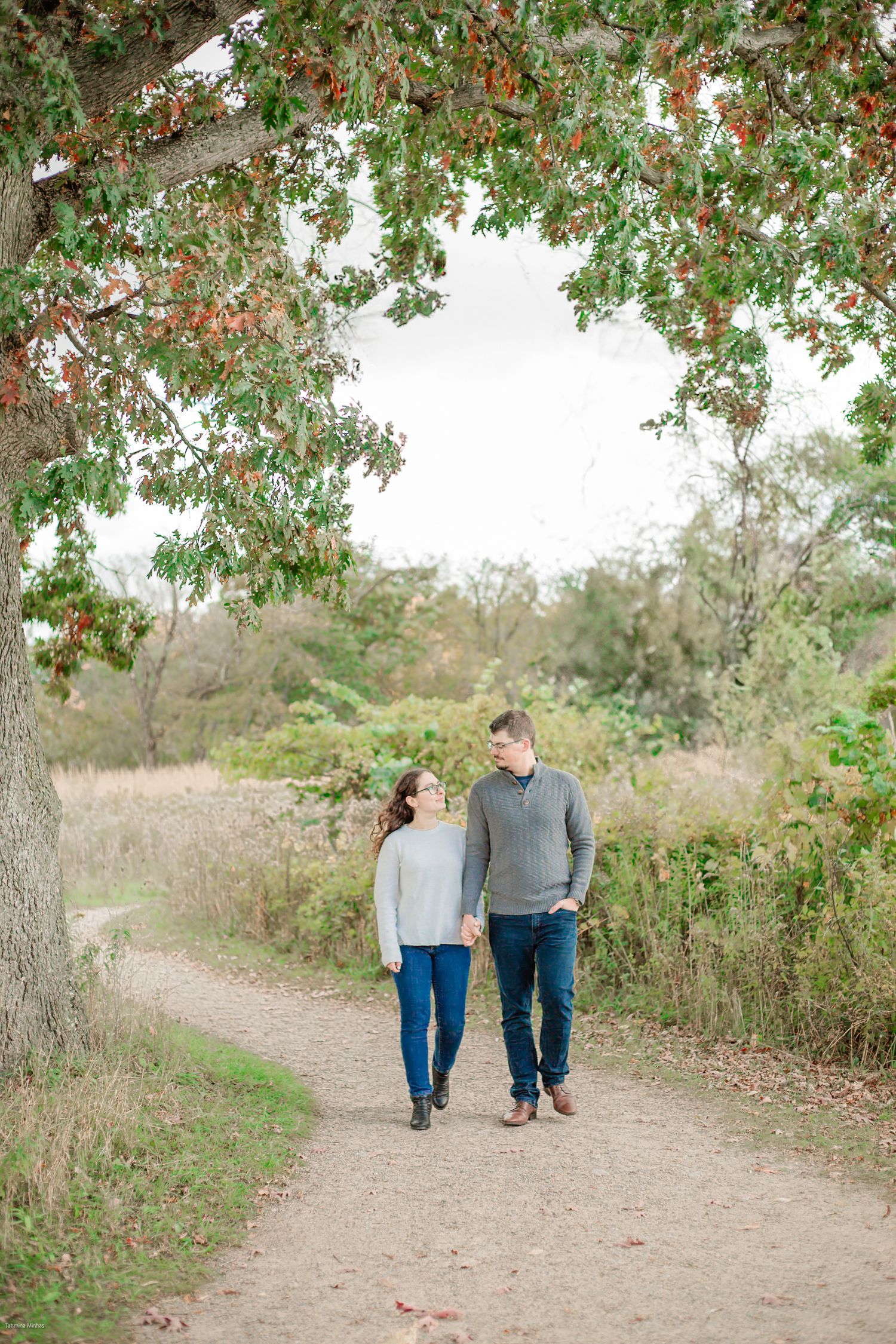 Two people walk hand in hand along a winding dirt path surrounded by lush greenery and trees.