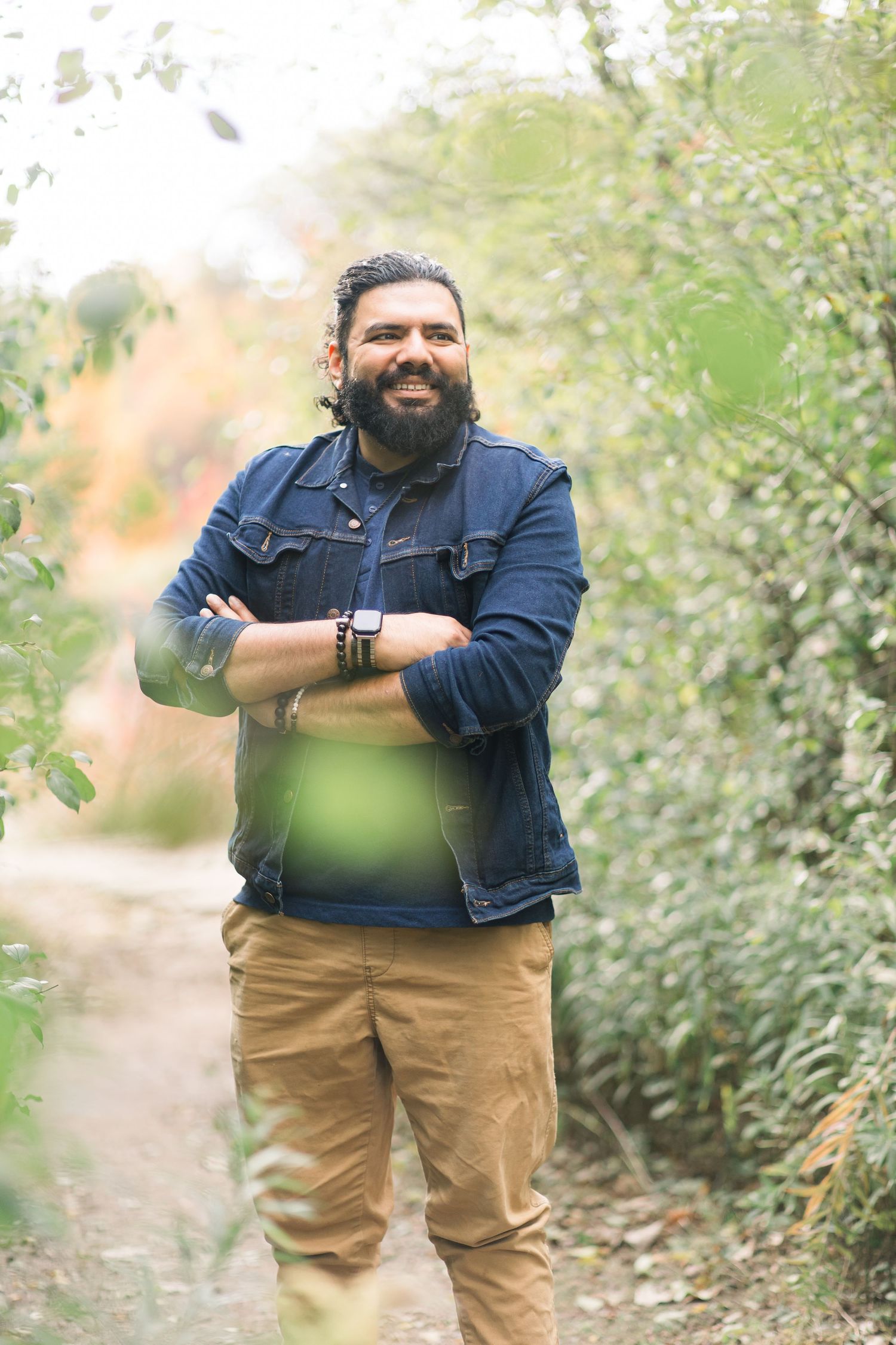 Person in denim jacket and khaki pants poses outdoors in natural setting with green foliage in background.