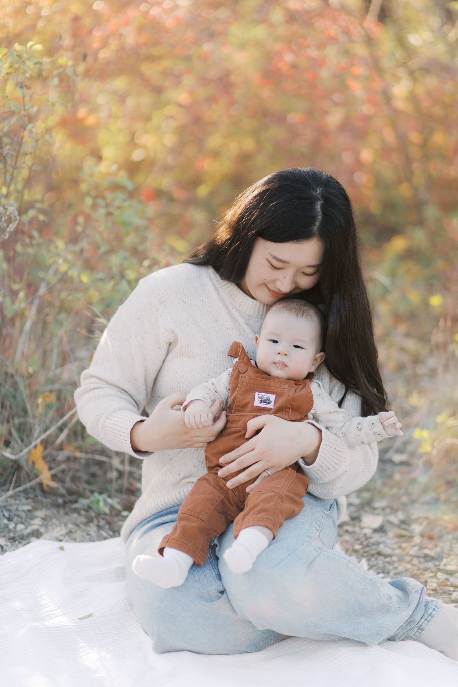 A mother in a beige sweater lovingly embraces and kisses her baby on a cozy blanket surrounded by autumn foliage.