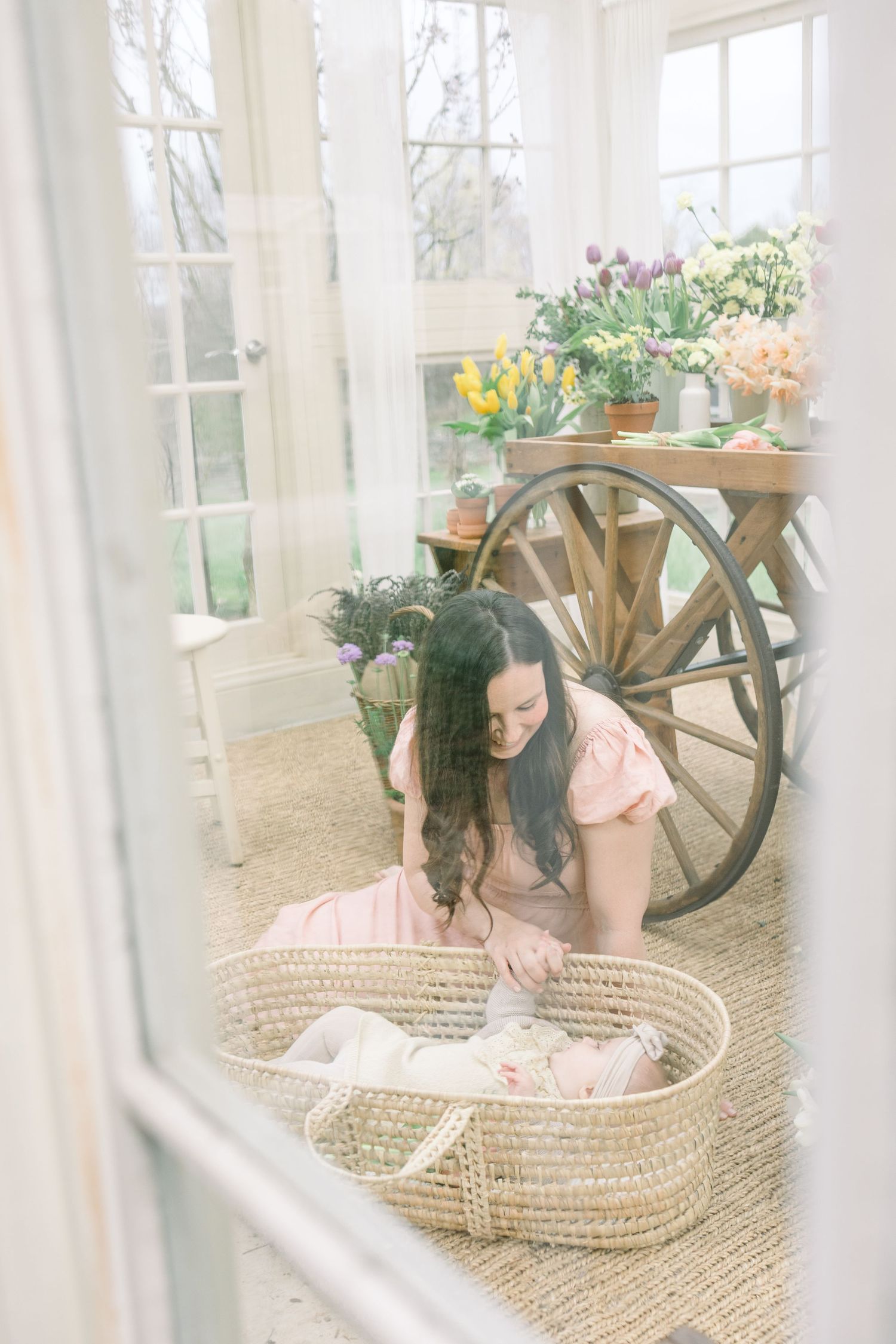 A woman leans over a woven baby bassinet in a bright sunlit room with wagon wheel decor and potted plants.