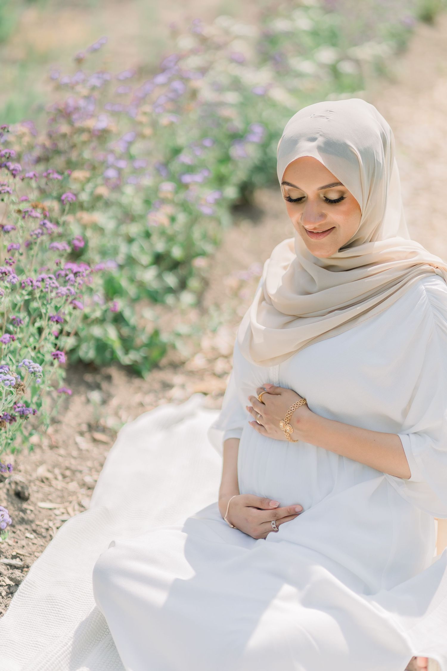 A woman in a white dress and beige hijab poses gracefully in a garden with purple wildflowers.