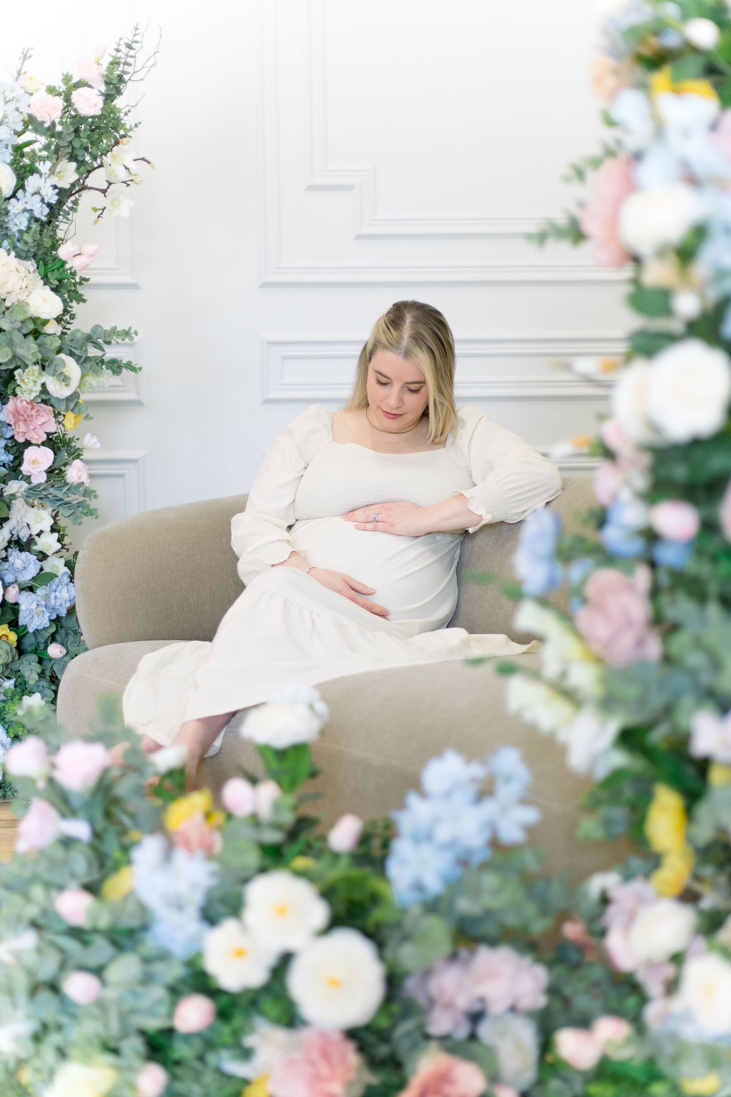 A maternity photoshoot featuring soft floral arrangements and white decor in an elegant indoor setting.