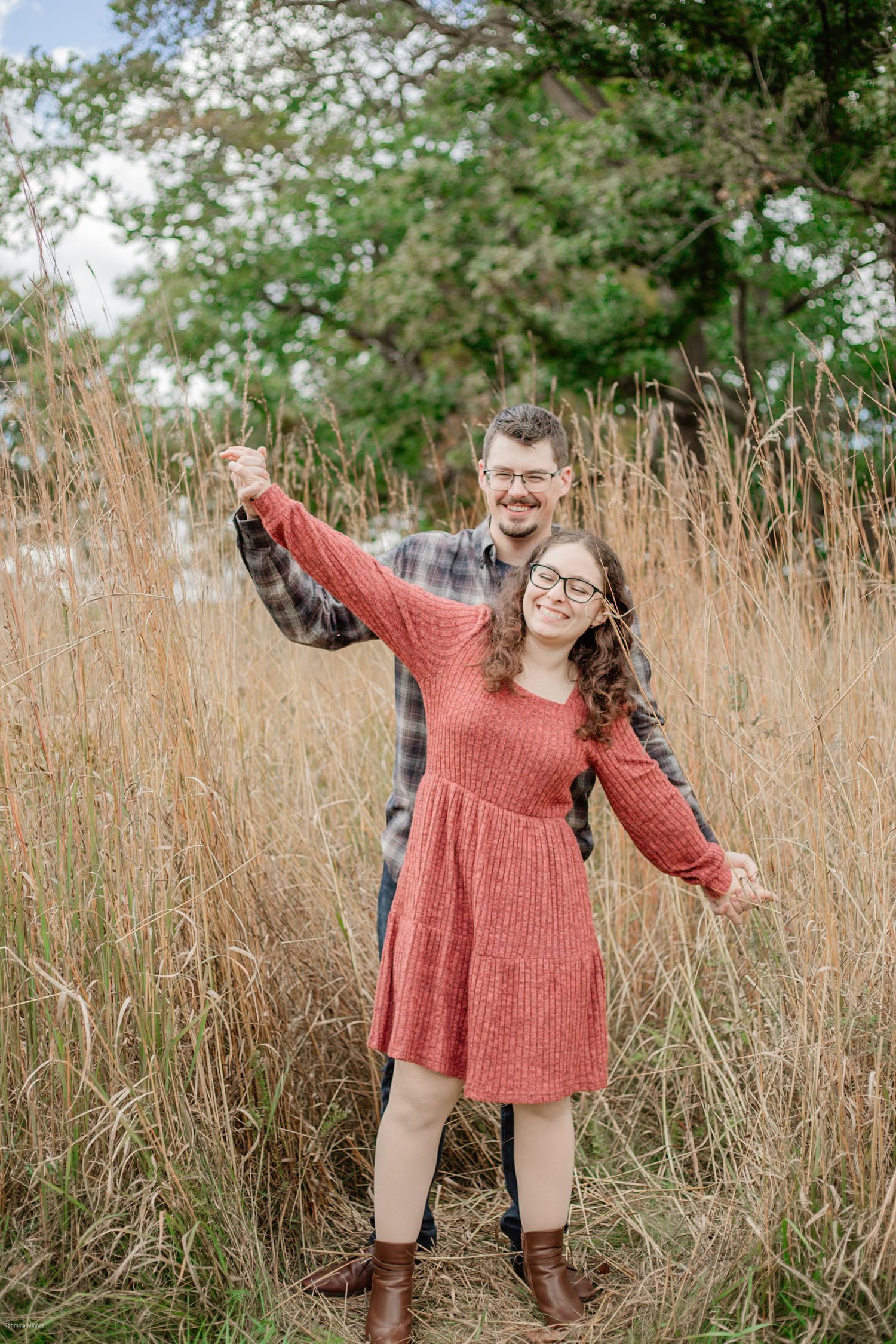 A couple stands playfully in a field of tall brown grass on an autumn day, with green trees visible in the background.
