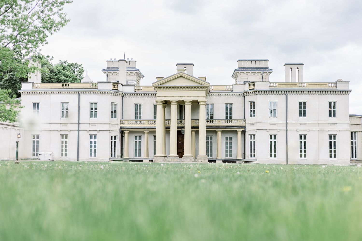 A grand white neoclassical mansion with columned entrance and symmetrical wings sits behind a lush green lawn.