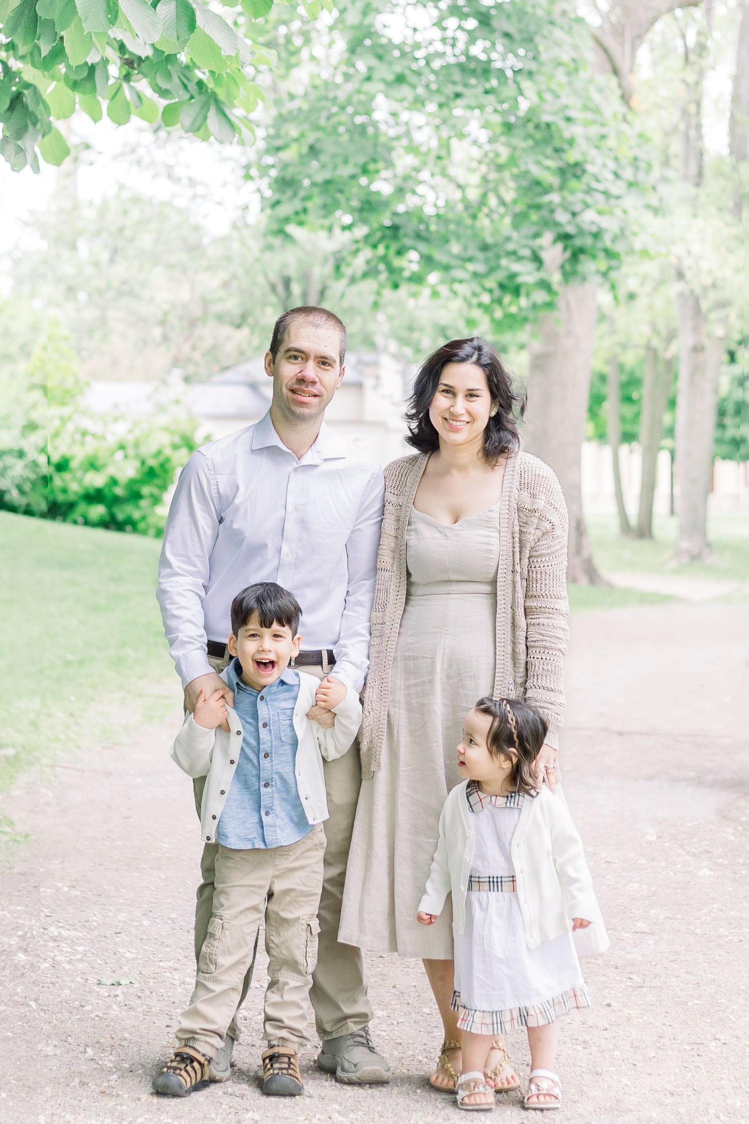 A family enjoys a casual outdoor photo session together in a serene park with green trees in the background.