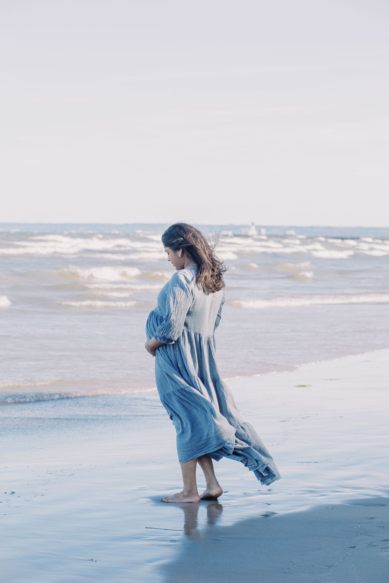 A person in a flowing blue dress walks along the shoreline of a beach at sunset.