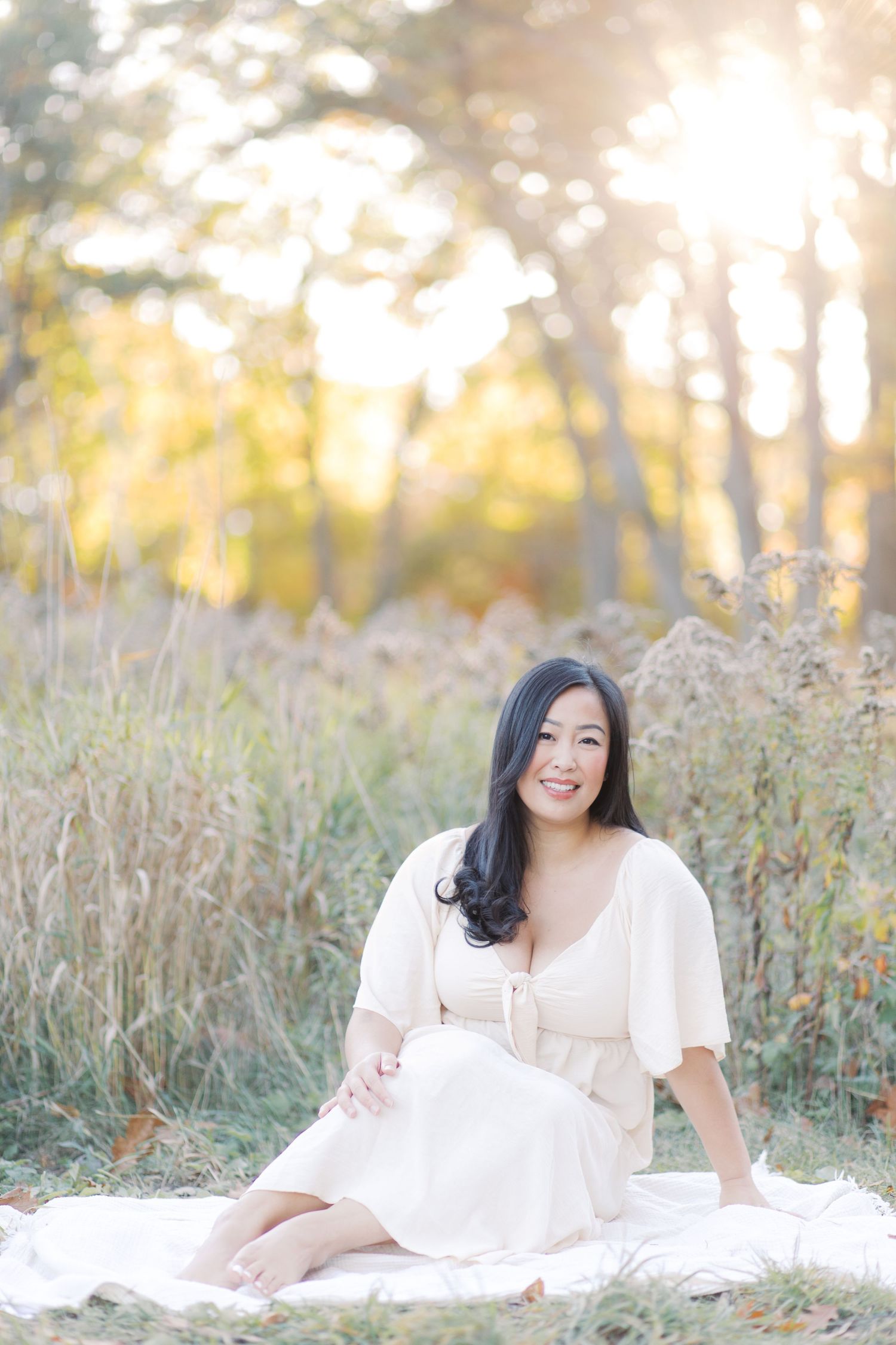 A person in a white dress lies on a blanket in a sunlit meadow with tall grass during golden hour.
