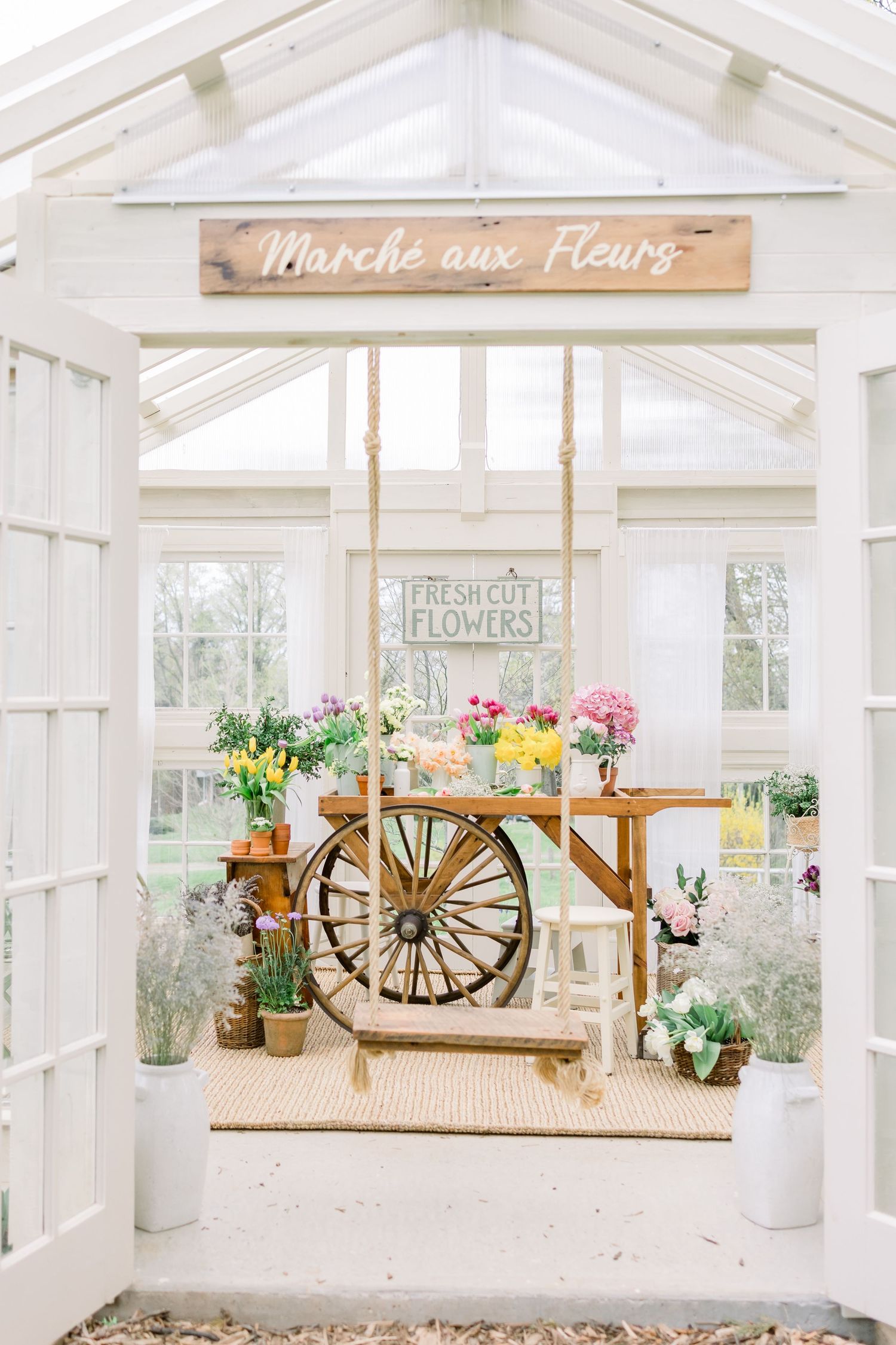 Bright white greenhouse interior with rustic wooden wagon wheel decor and potted flowers on display in a vintage farmhouse style.
