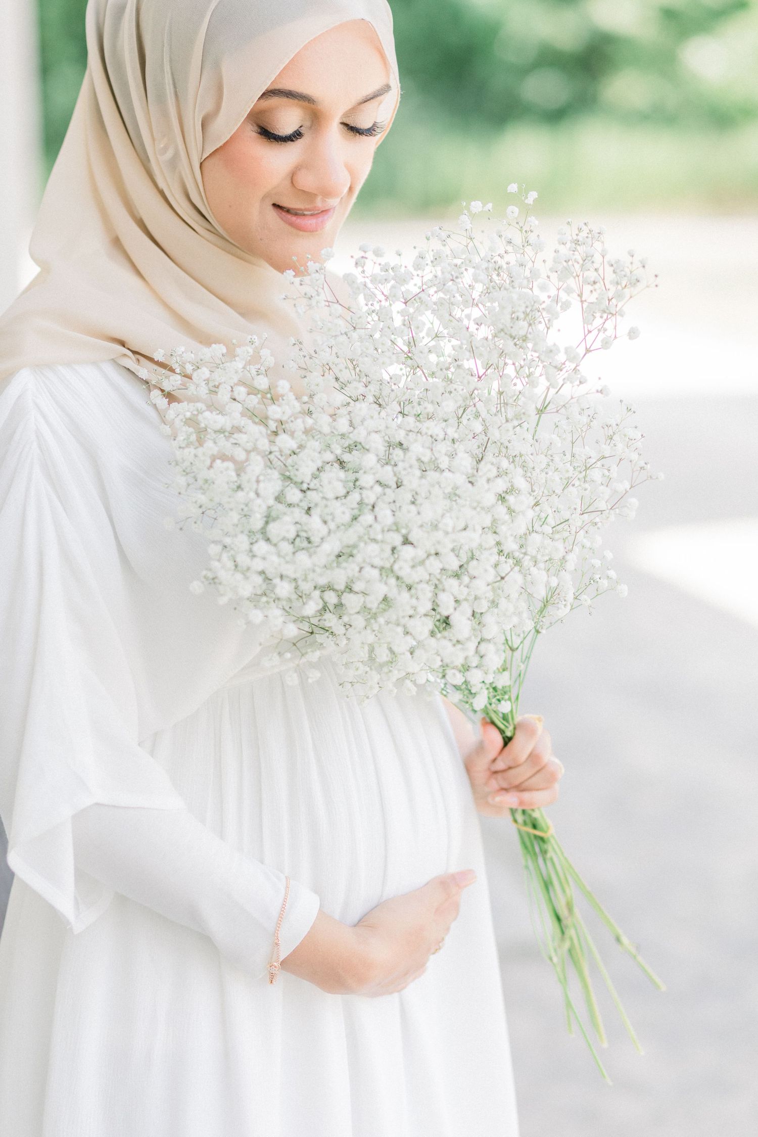 A person in white hijab and dress holds a delicate bouquet of baby's breath flowers in a bright outdoor setting.