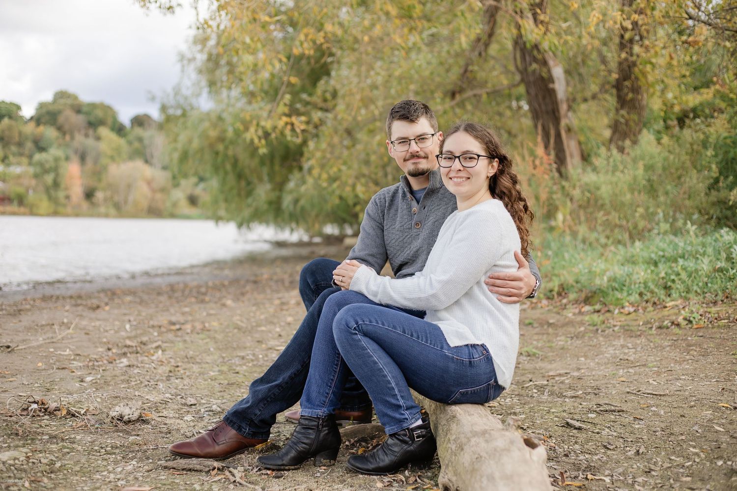 A couple sits together on a dirt path surrounded by autumn trees and foliage during an outdoor photo session.