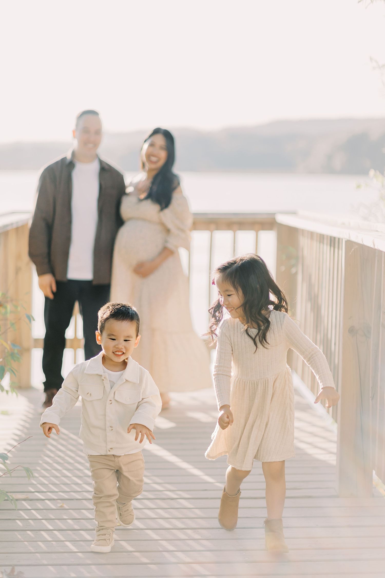 Children playing and running together on a sunlit wooden deck by the water on a bright afternoon.