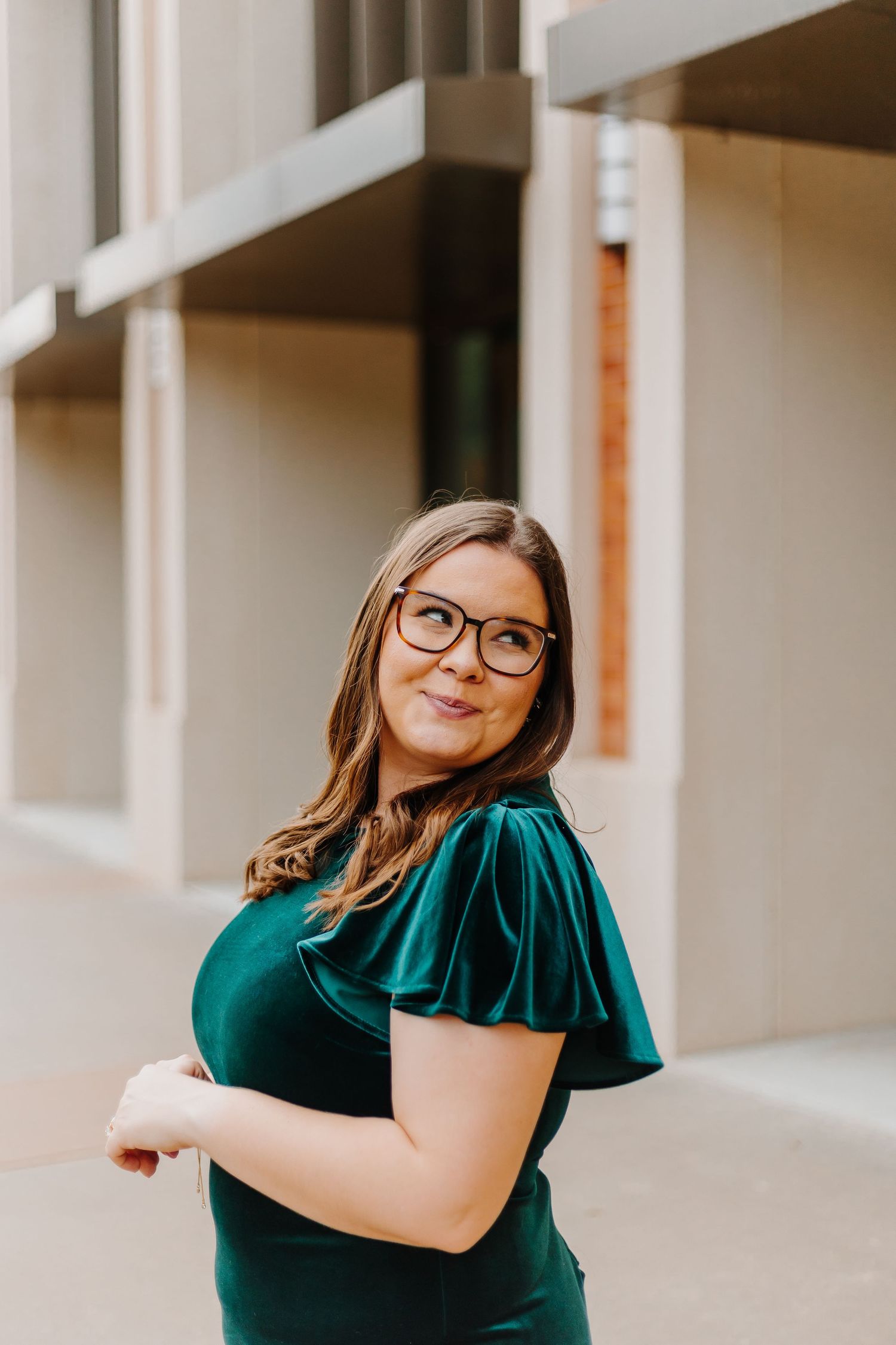 Woman in emerald green velvet dress and glasses poses against light colored wall in outdoor portrait series.