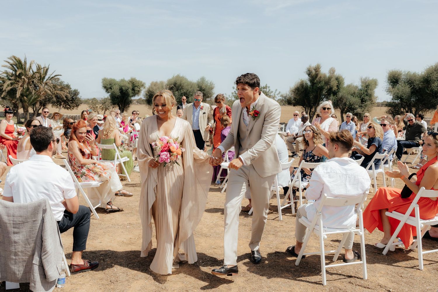Wedding guests celebrate with newlyweds during an outdoor ceremony with palm trees in the background.