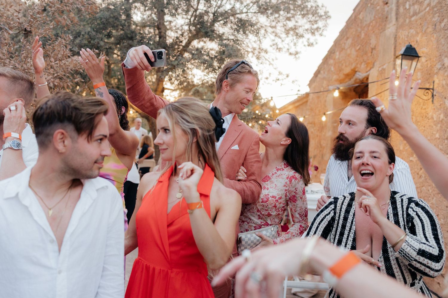 A joyous crowd celebrates and dances together at an outdoor party with string lights visible.