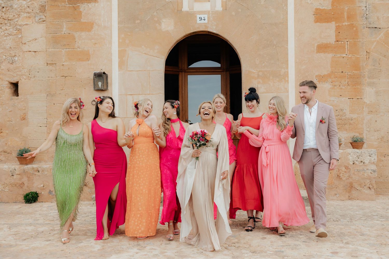 Wedding party wearing colorful dresses in different shades of pink, red, green and orange poses against stone building.