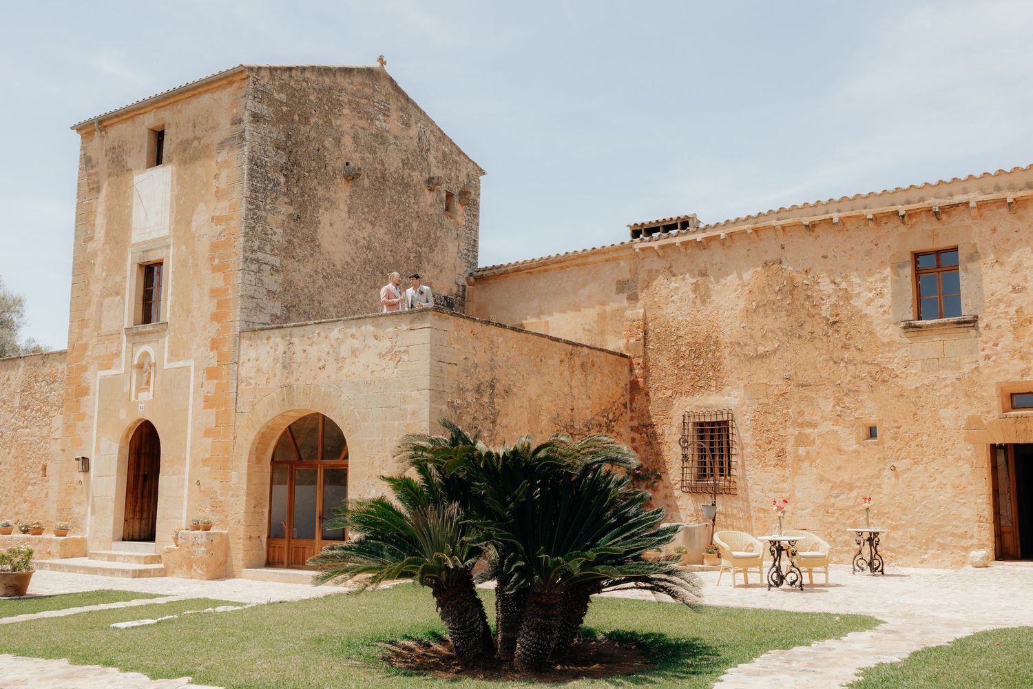 Mediterranean stone villa with rustic tower entrance, arched doorway, and palm tree in courtyard outside historic building.