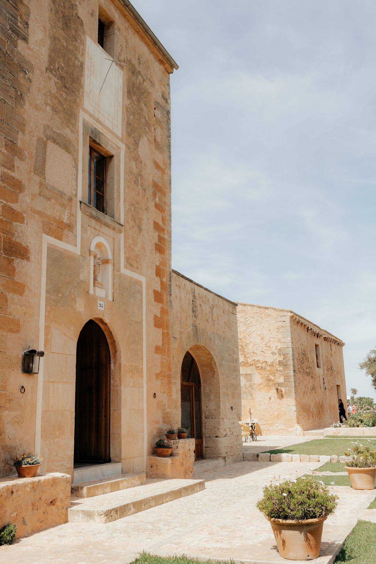 Ancient Mediterranean stone building exterior with arched doorways and potted plants along a paved pathway.