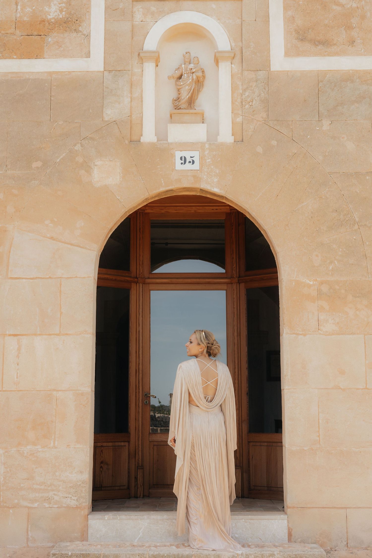 Elegant backless dress silhouette against rustic stone archway with statue alcove above.