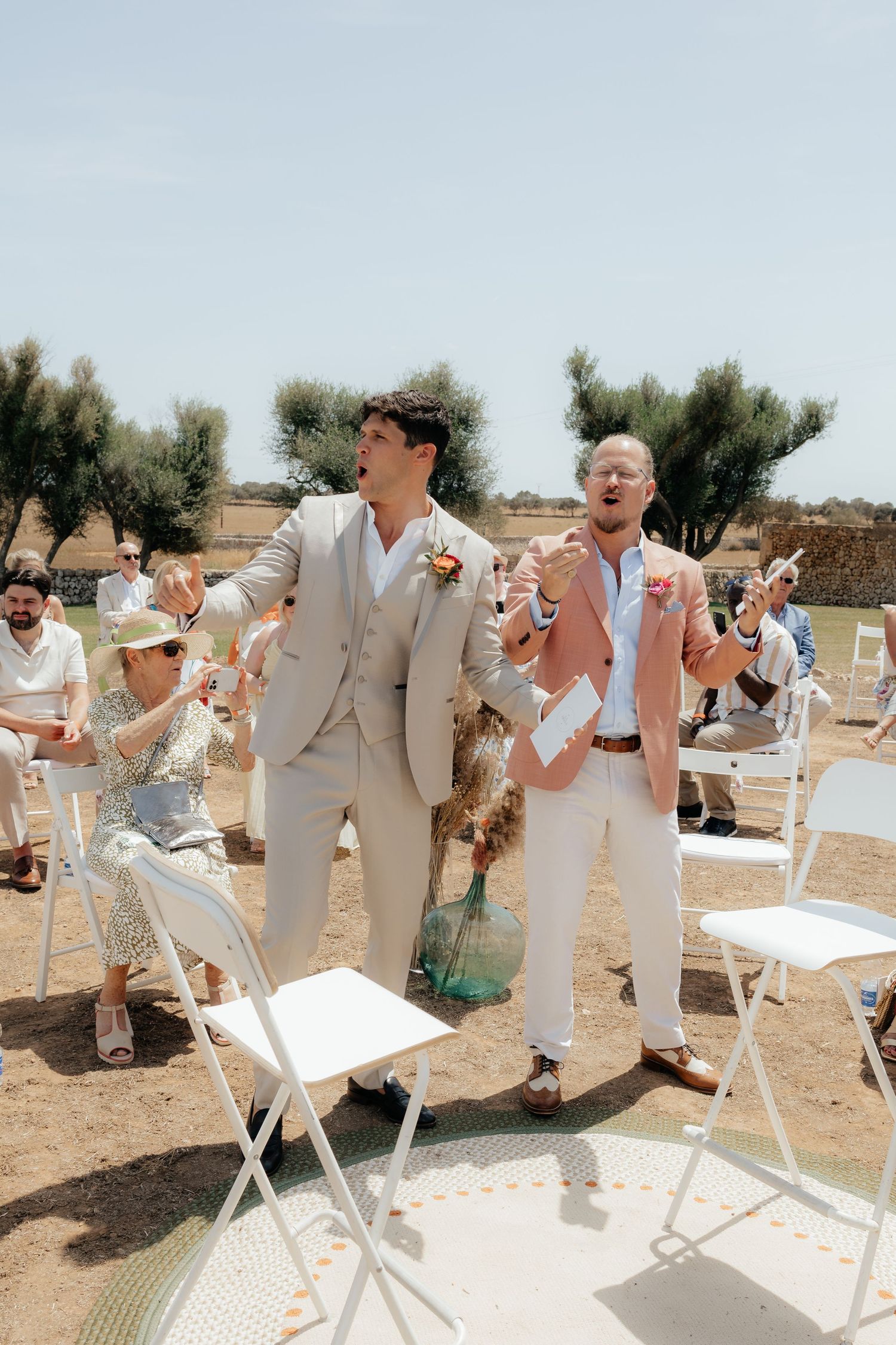 Wedding guests celebrating joyfully at an outdoor ceremony with standing guests in light-colored attire.