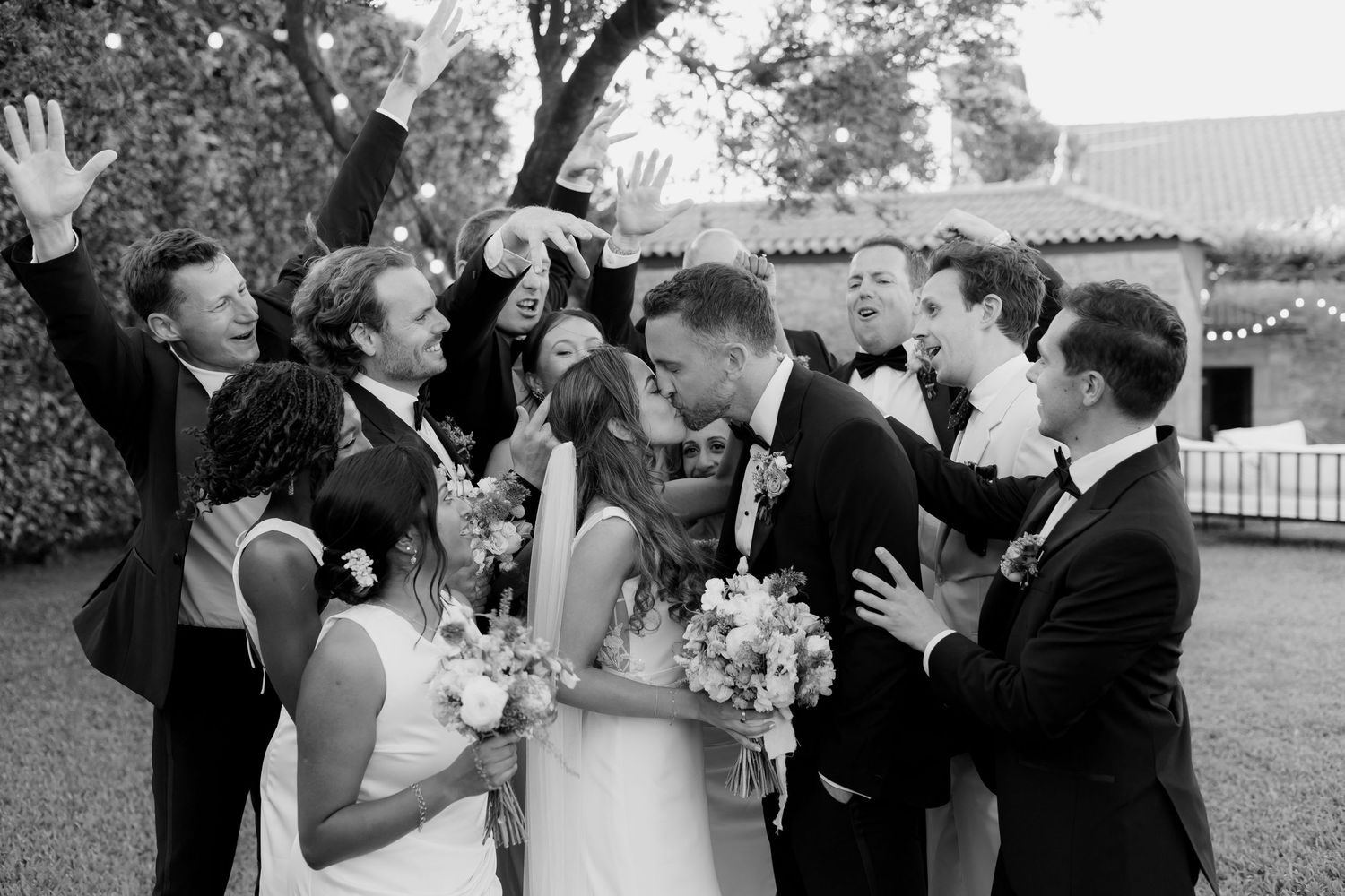 A joyful wedding party celebrates outdoors with raised hands and laughter in a black and white candid moment.