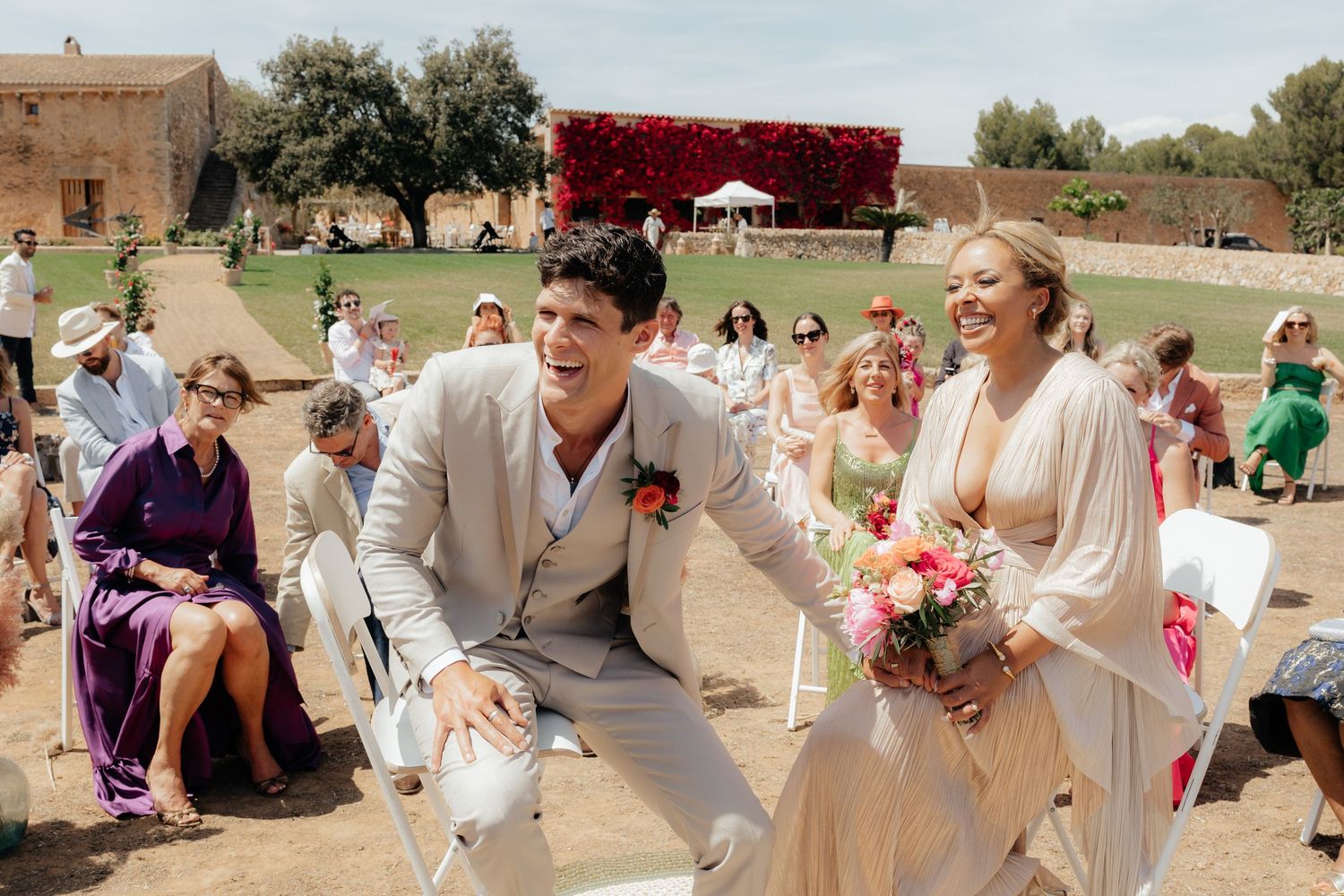Wedding celebration at a rustic outdoor venue with guests seated on white chairs and vibrant red floral decor.