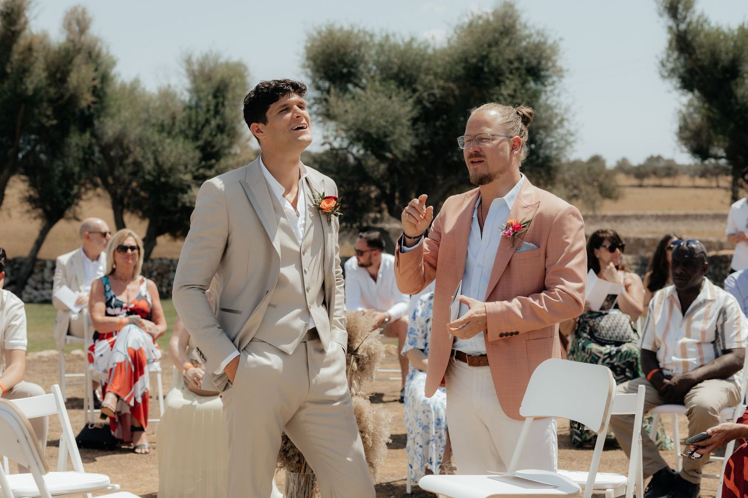 Two people in light summer suits share a laugh during an outdoor event with white folding chairs and guests in background.