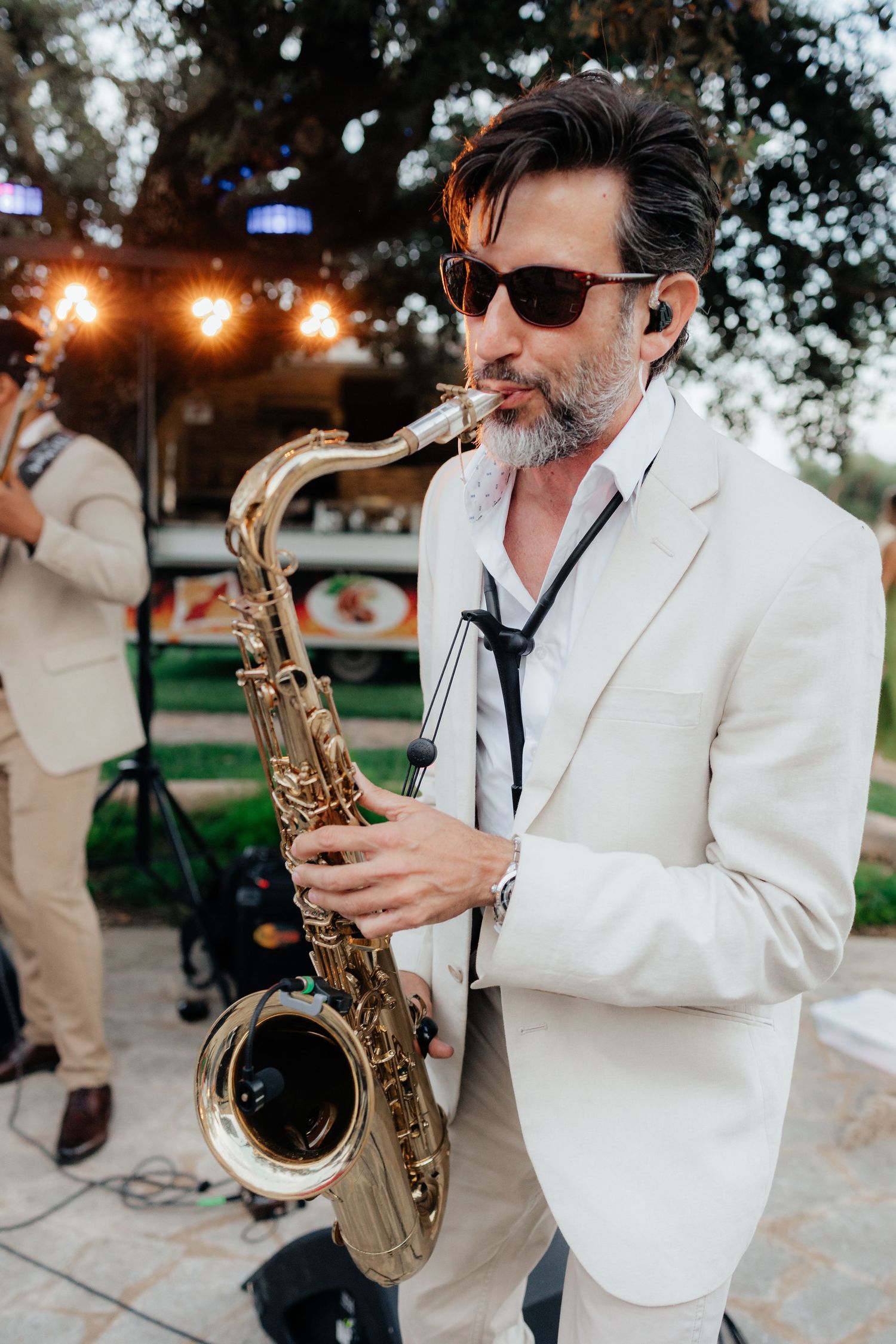 Saxophone player in white blazer and sunglasses performing at outdoor event.