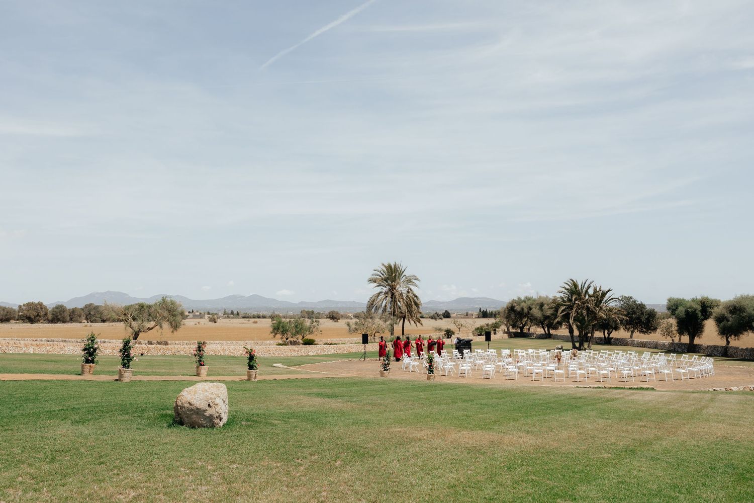 An outdoor wedding ceremony setup with white chairs on a grassy area at a rustic venue with palm trees.