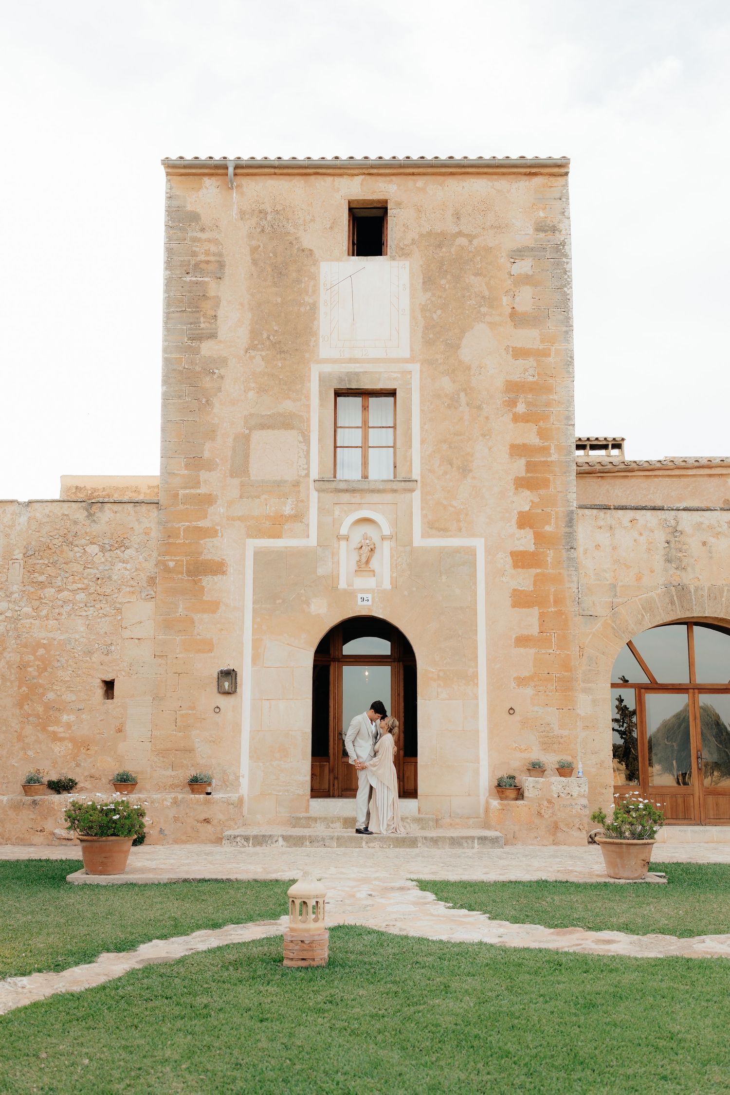 A romantic Mediterranean villa entrance with stone walls and a manicured lawn path leading to an arched doorway.