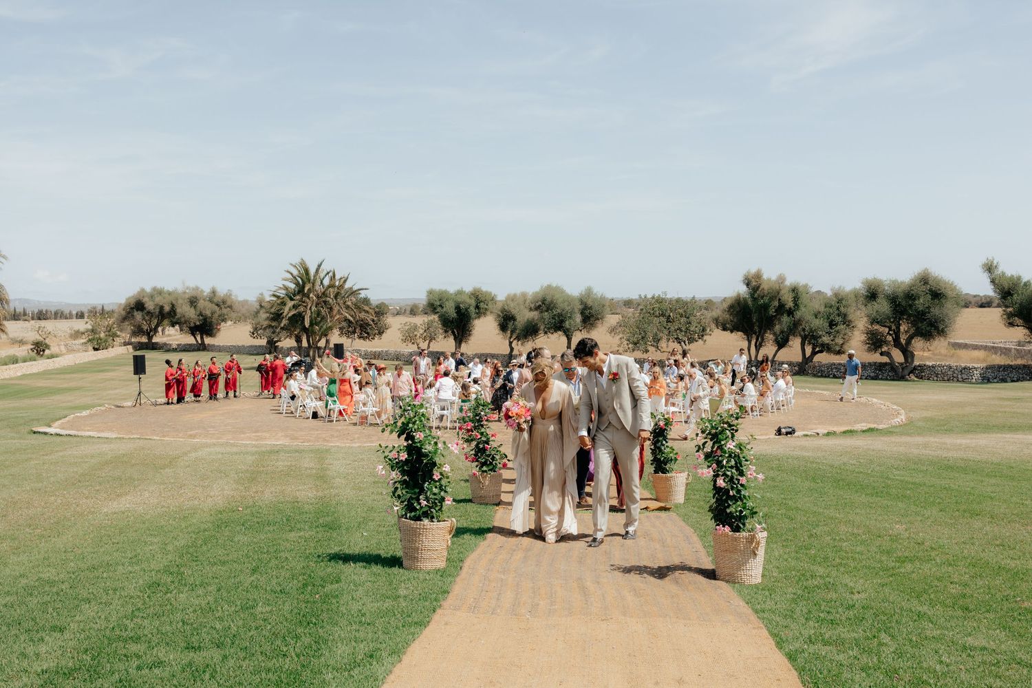 Wedding party walking down an outdoor path lined with greenery and guests.