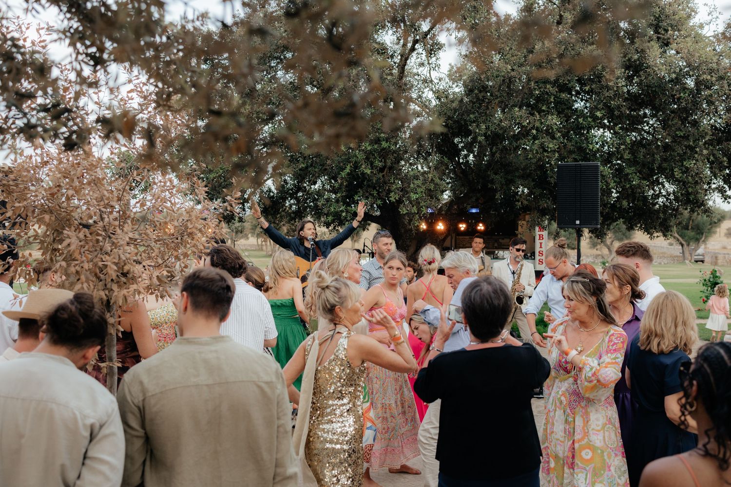 A lively outdoor celebration with guests dancing under string lights and trees at dusk.