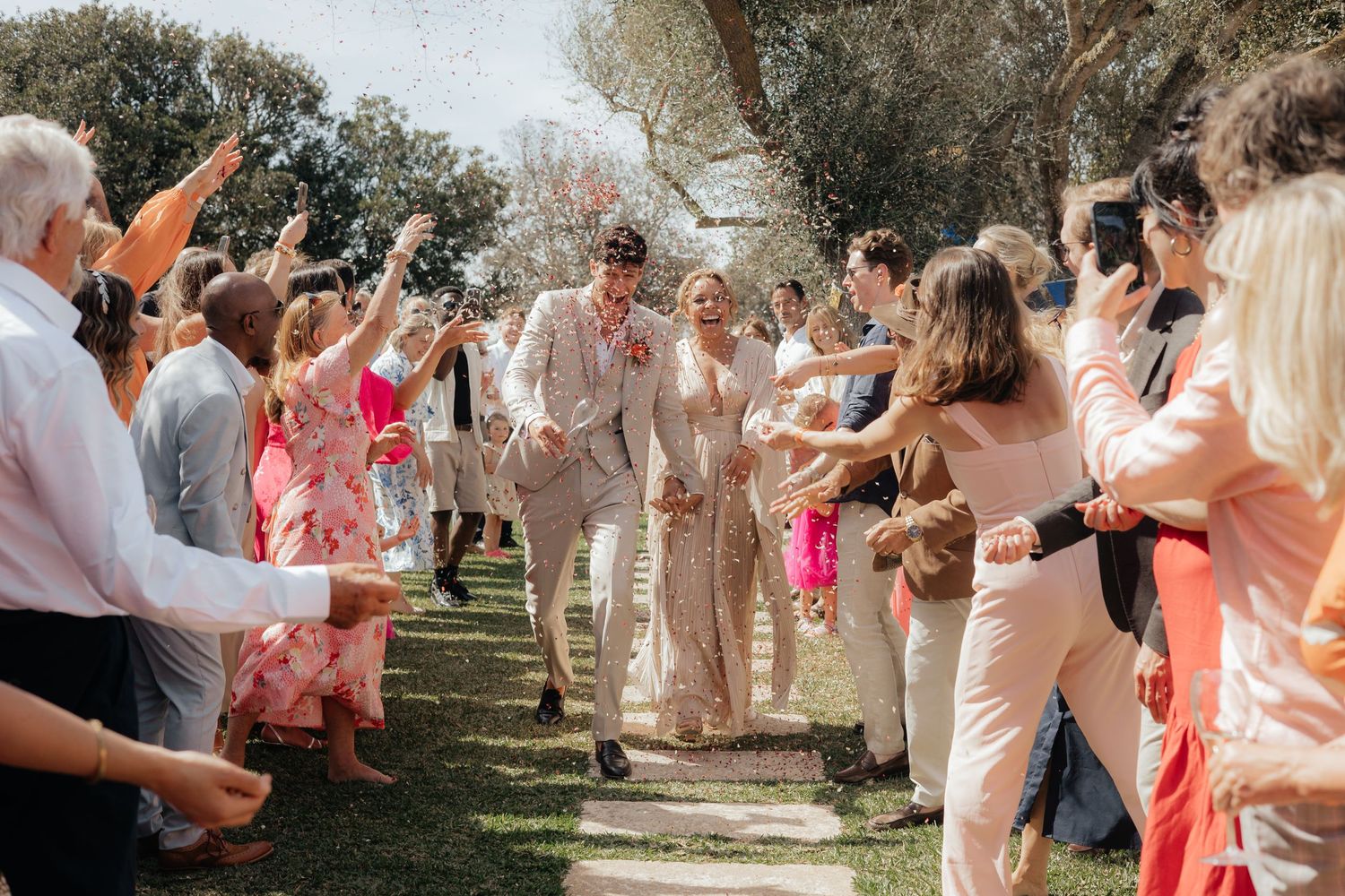 Wedding guests throw confetti and cheer as couple walks down outdoor aisle lined with trees.