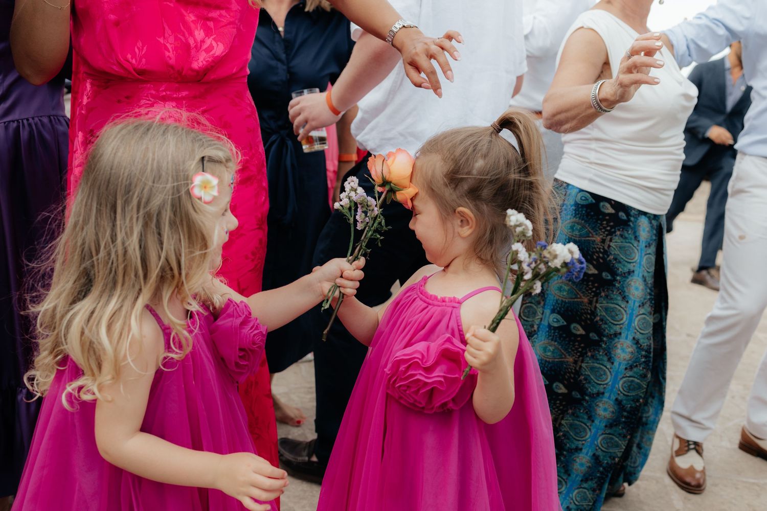 Two young children in matching bright pink dresses interact at an outdoor gathering.