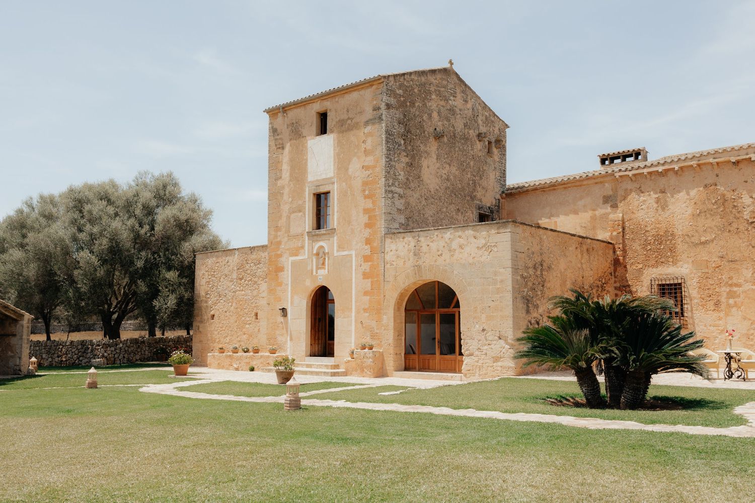 Ancient stone building with arched entrance and tower facade set on manicured lawn with olive trees in Mediterranean setting.