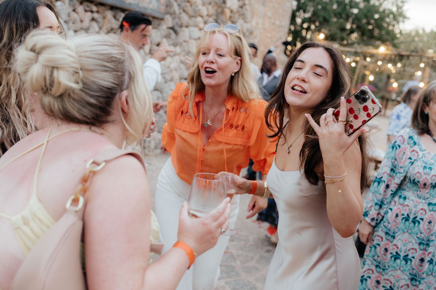 Friends enjoying drinks and laughter during outdoor celebration with string lights in background.