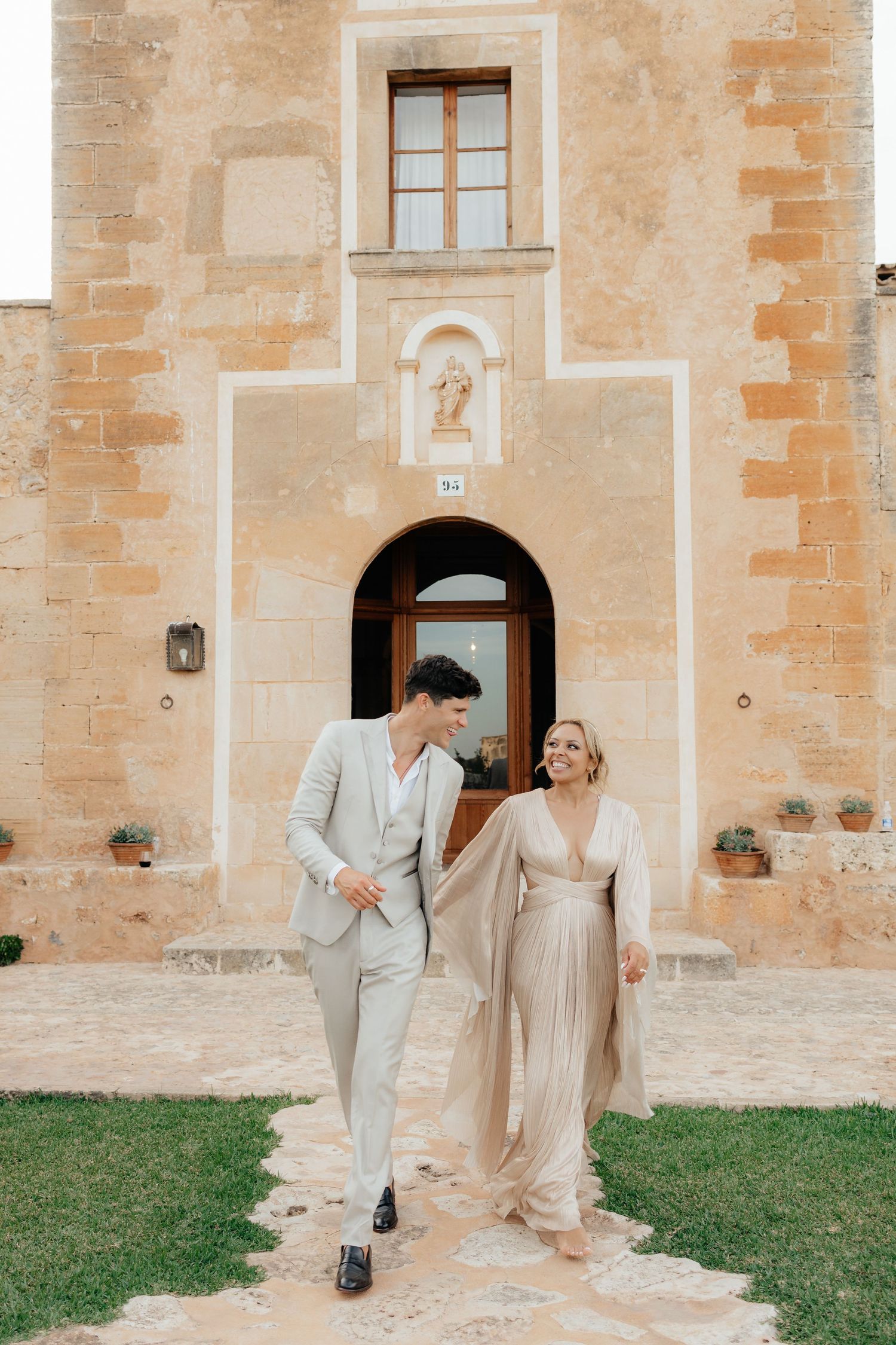 Elegant couple embraces in front of a rustic Mediterranean stone building entrance with an arched doorway.