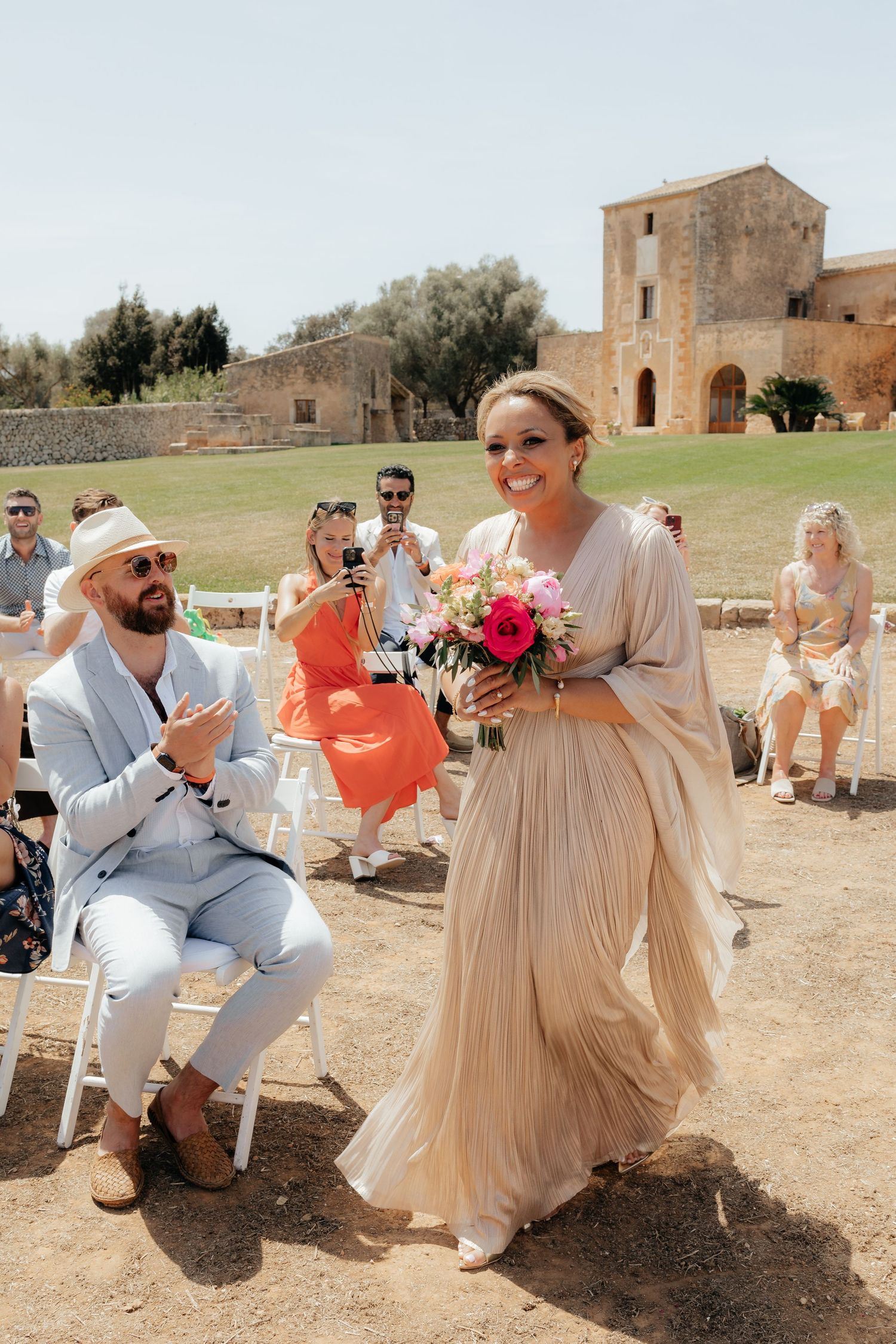 Wedding celebration at a rustic stone venue with guests seated on white chairs outdoors.