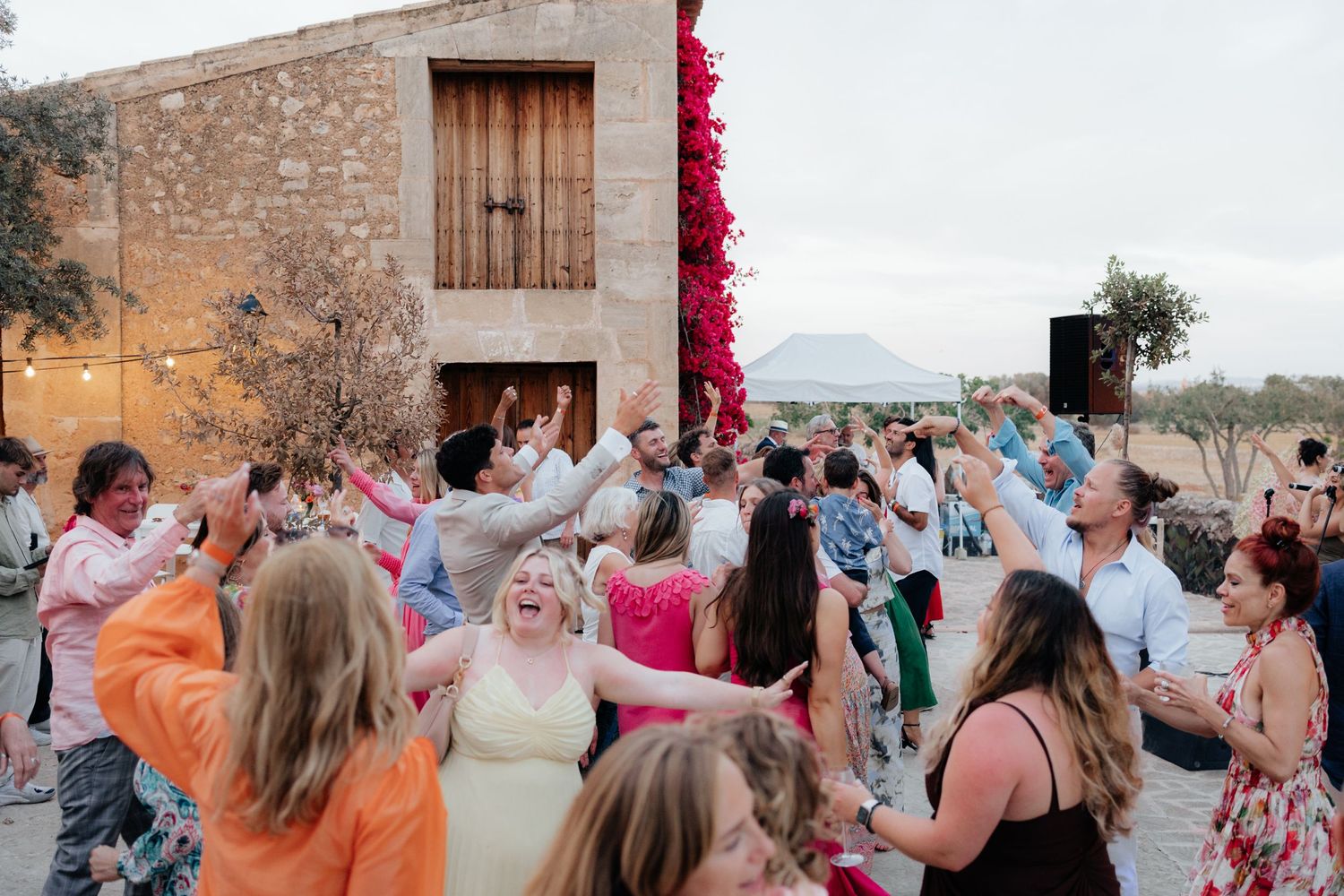 Large group of wedding guests dancing together at outdoor stone venue with mountains in background.
