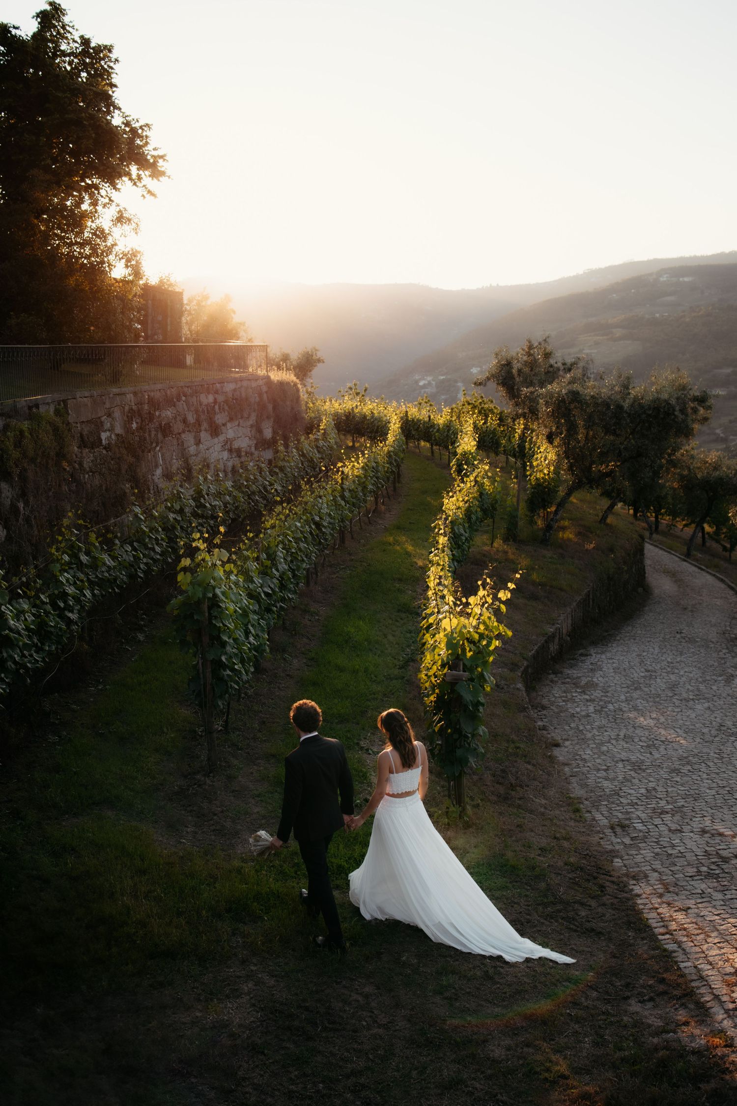 A couple stands in a scenic vineyard at sunset with rolling hills and stone walls in the background.