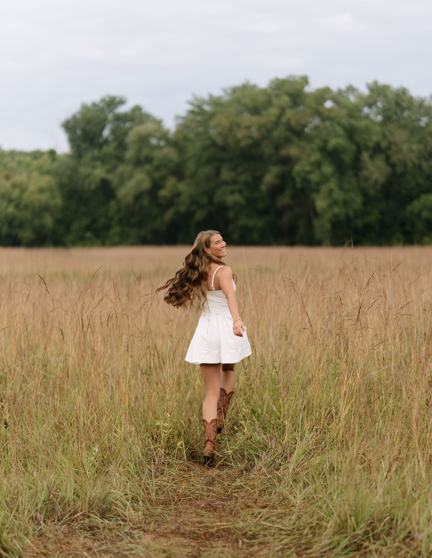 Girl with long wavy hair in a white dress and cowboy boots twirls in a tall grass meadow beneath an overcast sky.