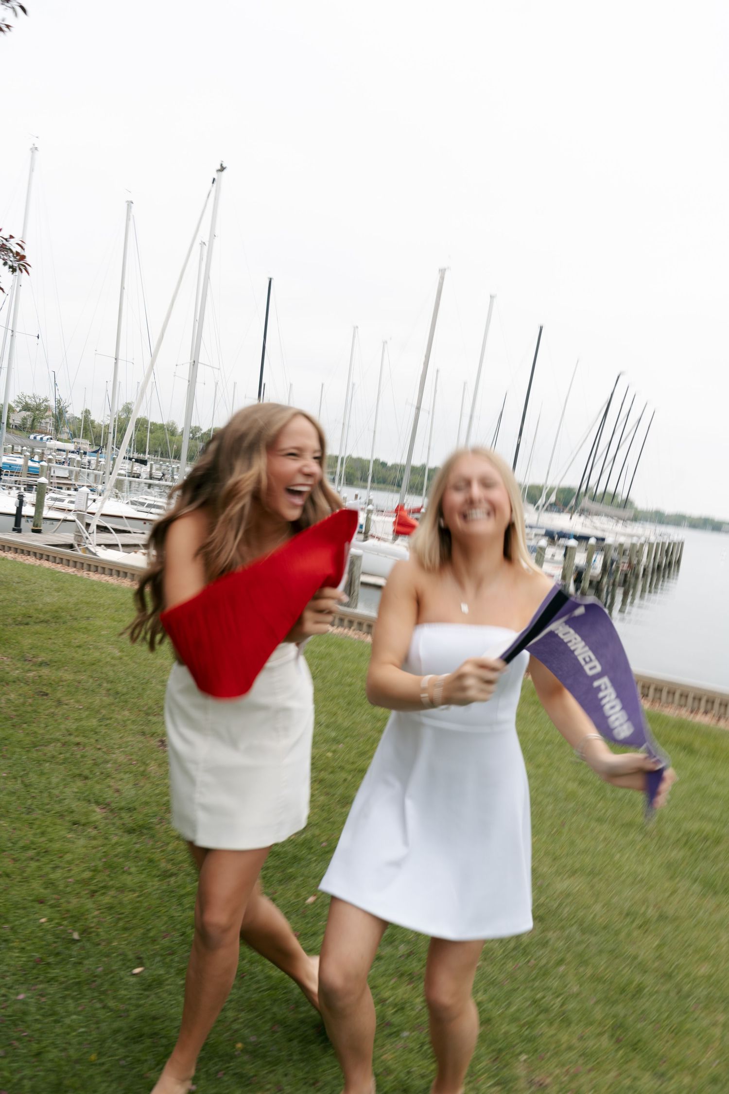Two laughing young women in white dresses play with red and purple pennant flags on a lawn beside a marina with sailboats.