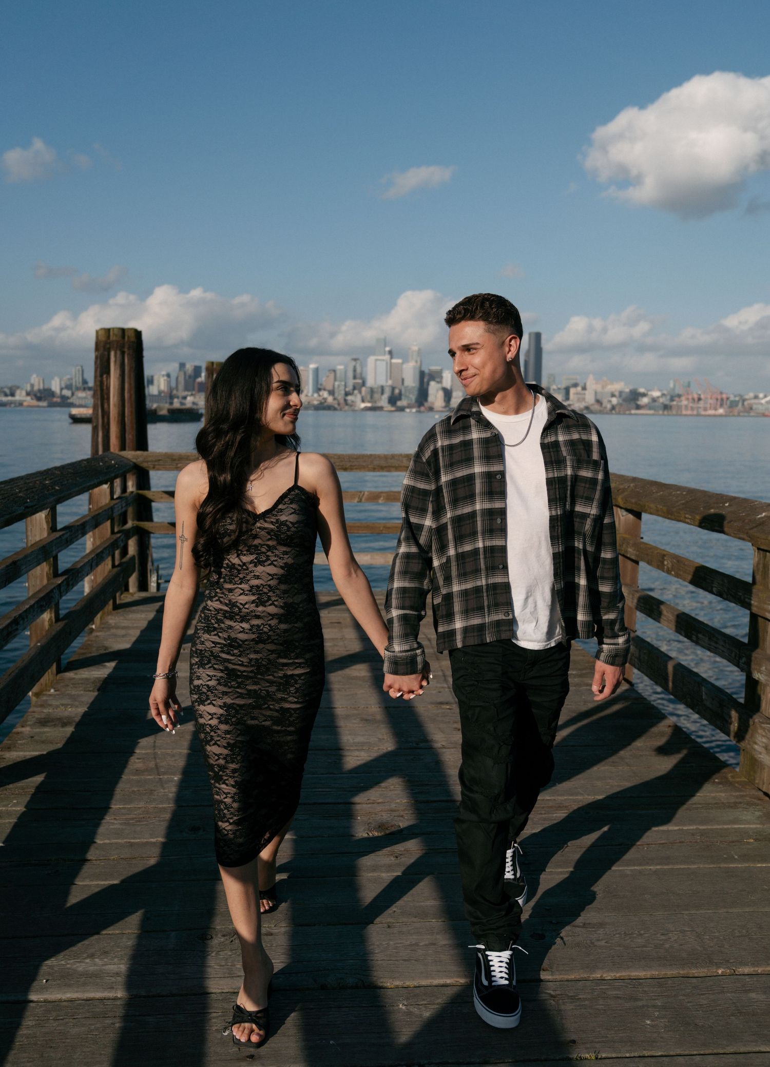 A couple holds hands and walks along a sunlit wooden pier with a city skyline across the water.