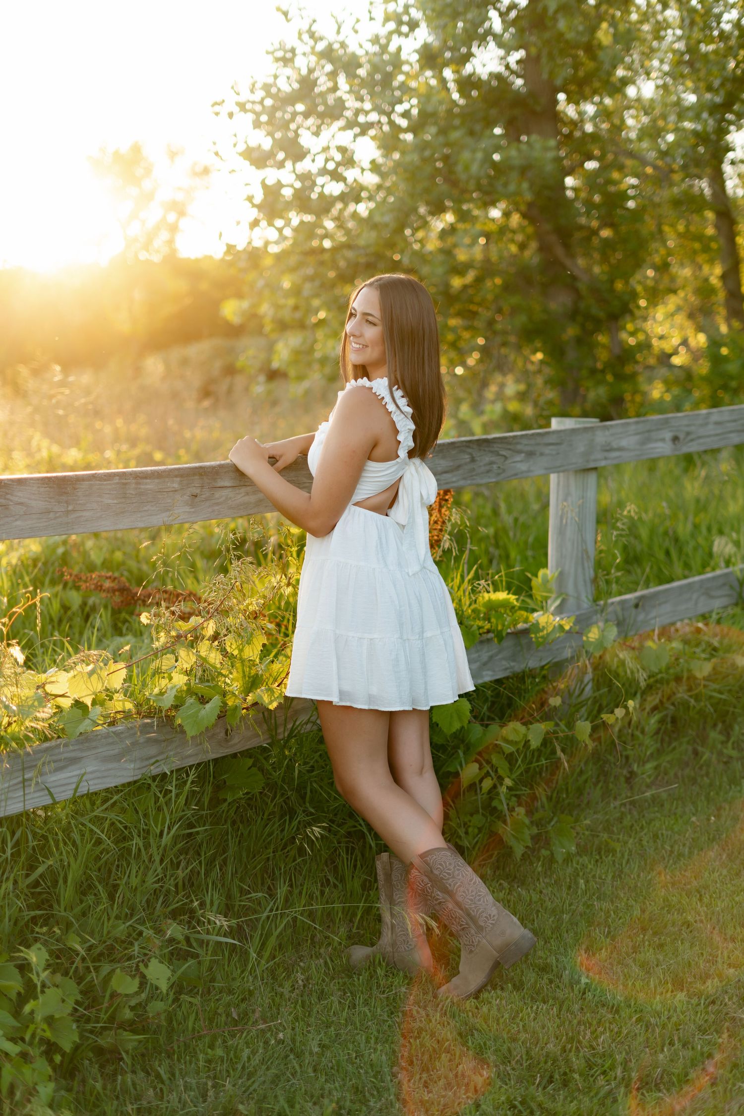 Young woman in a white sundress and cowboy boots leaning on a wooden fence in golden sunset light.
