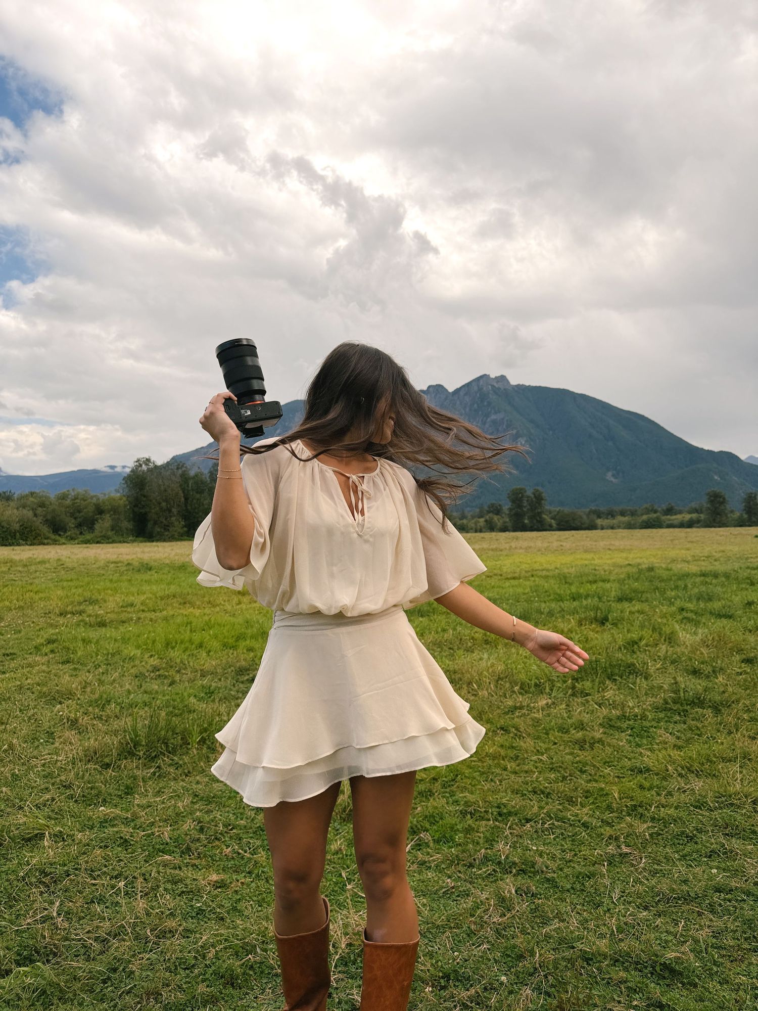 A woman in a cream ruffled dress and brown boots holds a camera while twirling in a grassy field with mountains and clouds behind.