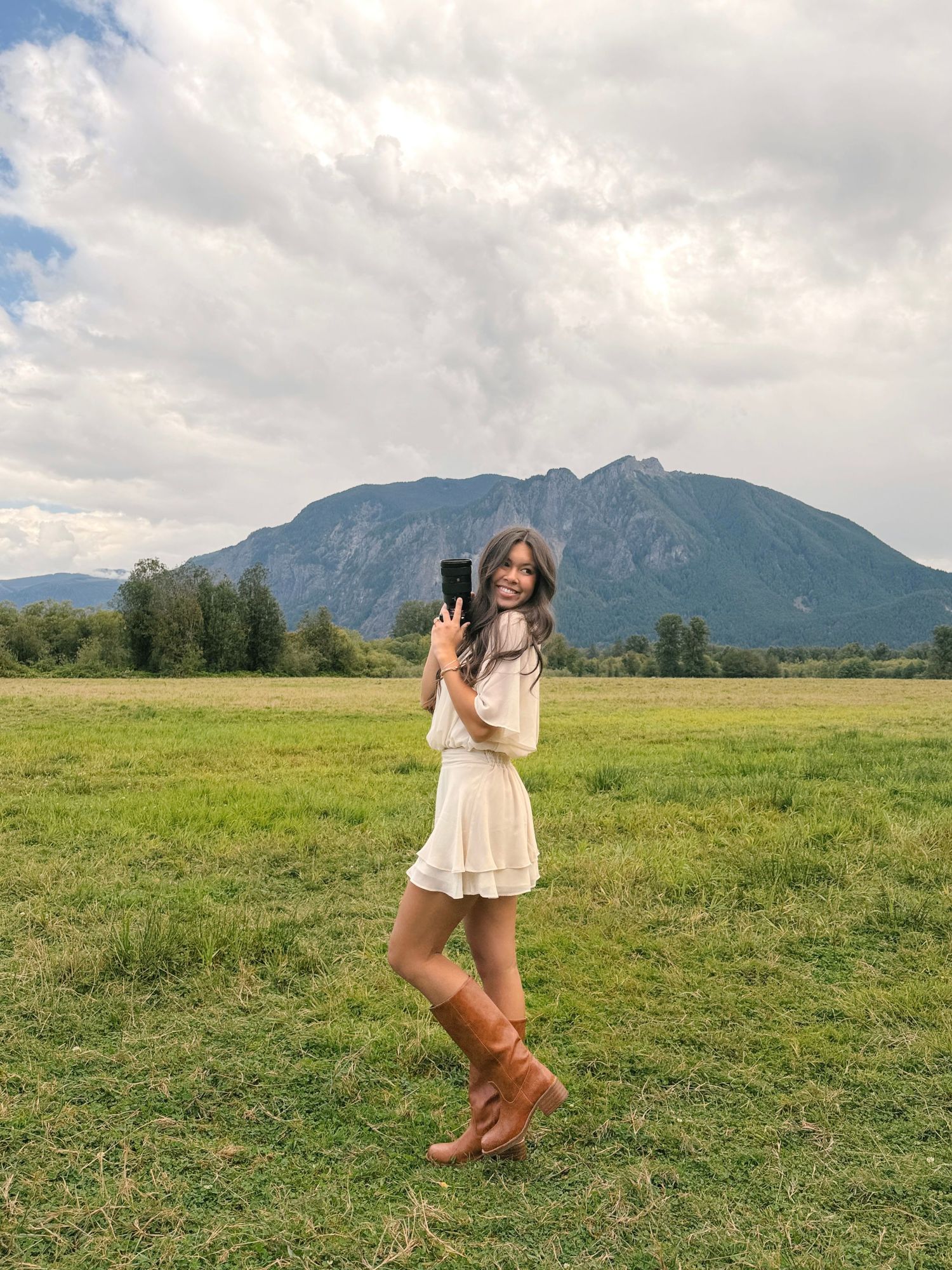Woman in a cream dress and tall brown boots takes a photo with her phone in a green field with a mountain behind her.