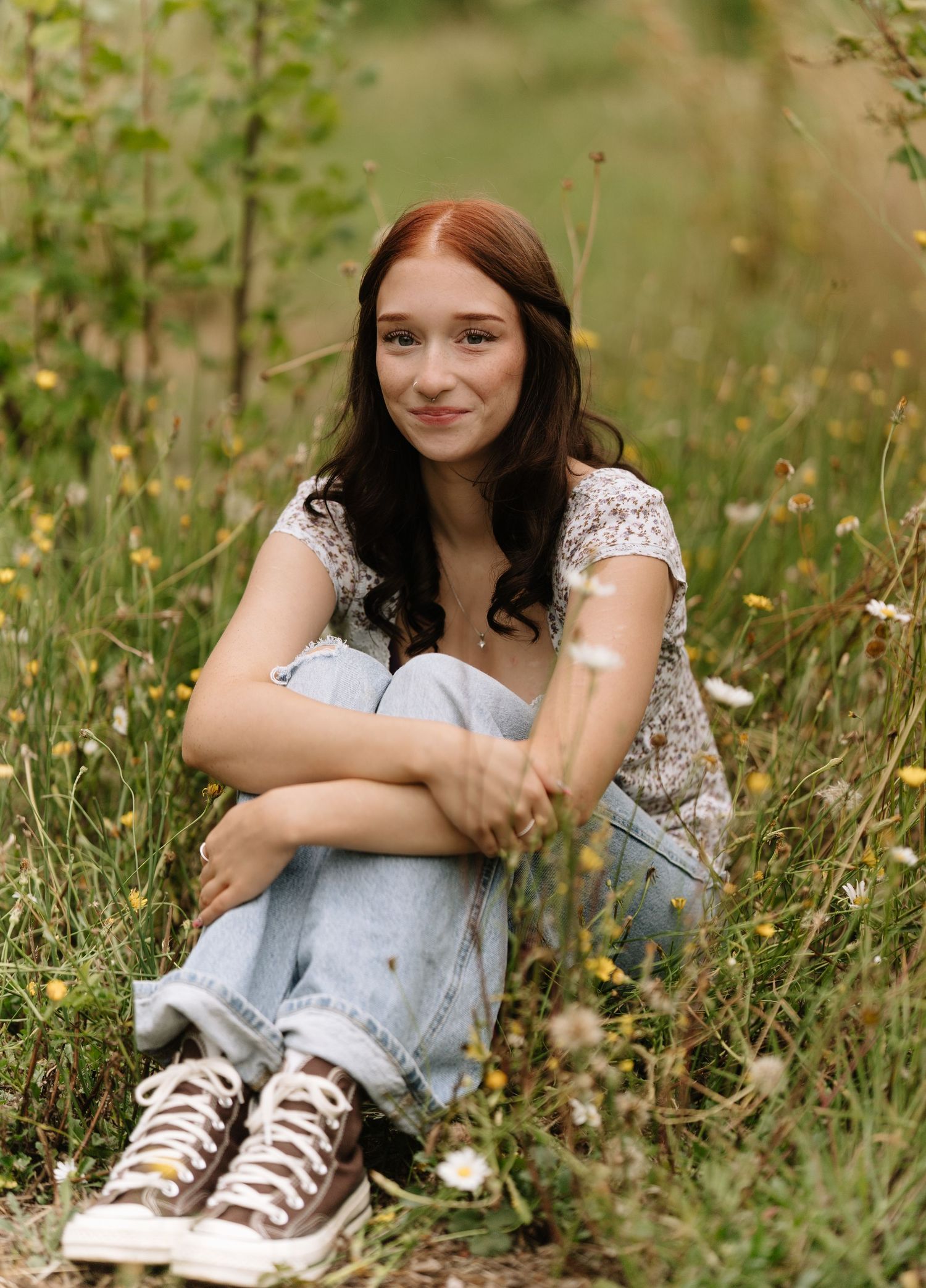 Young woman with auburn hair and denim jeans sits hugging her knees among wildflowers in a tall grass meadow, smiling softly.