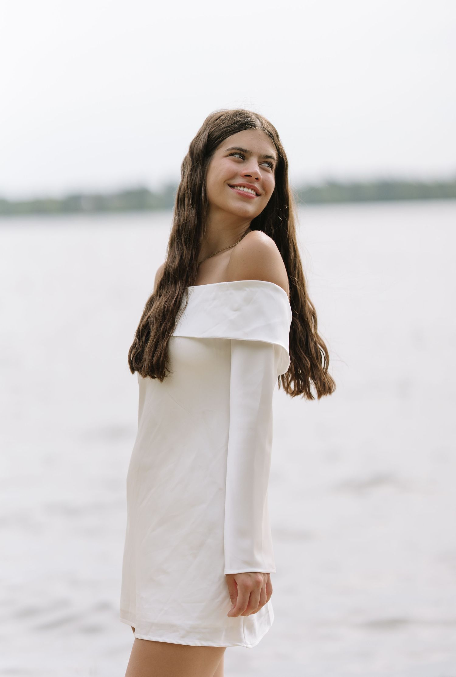 Young woman with long wavy hair wearing a white off-the-shoulder dress smiling by a calm lake.