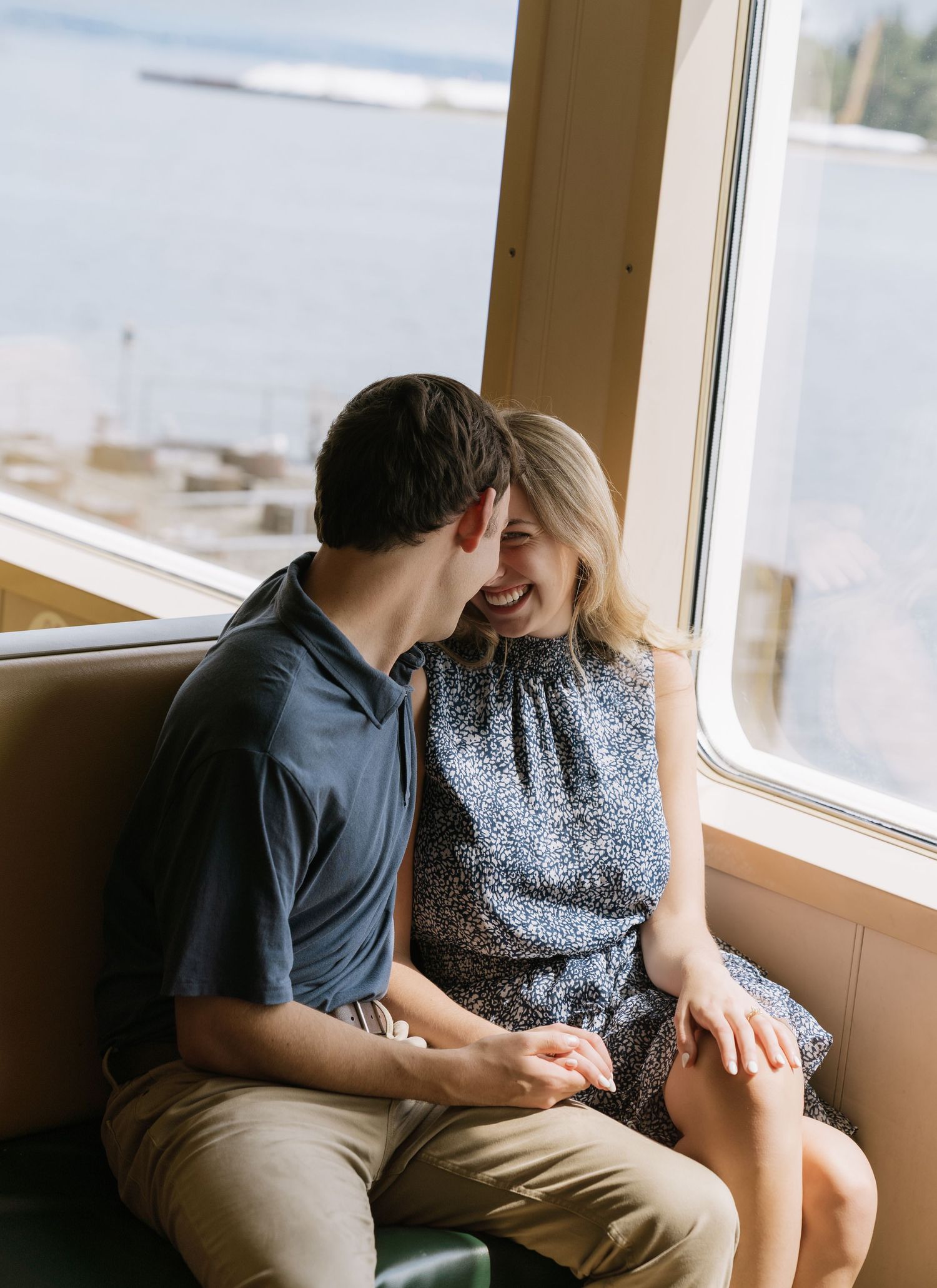 Couple laughs and cuddles on a bench by a window overlooking the water during a ferry engagement session.